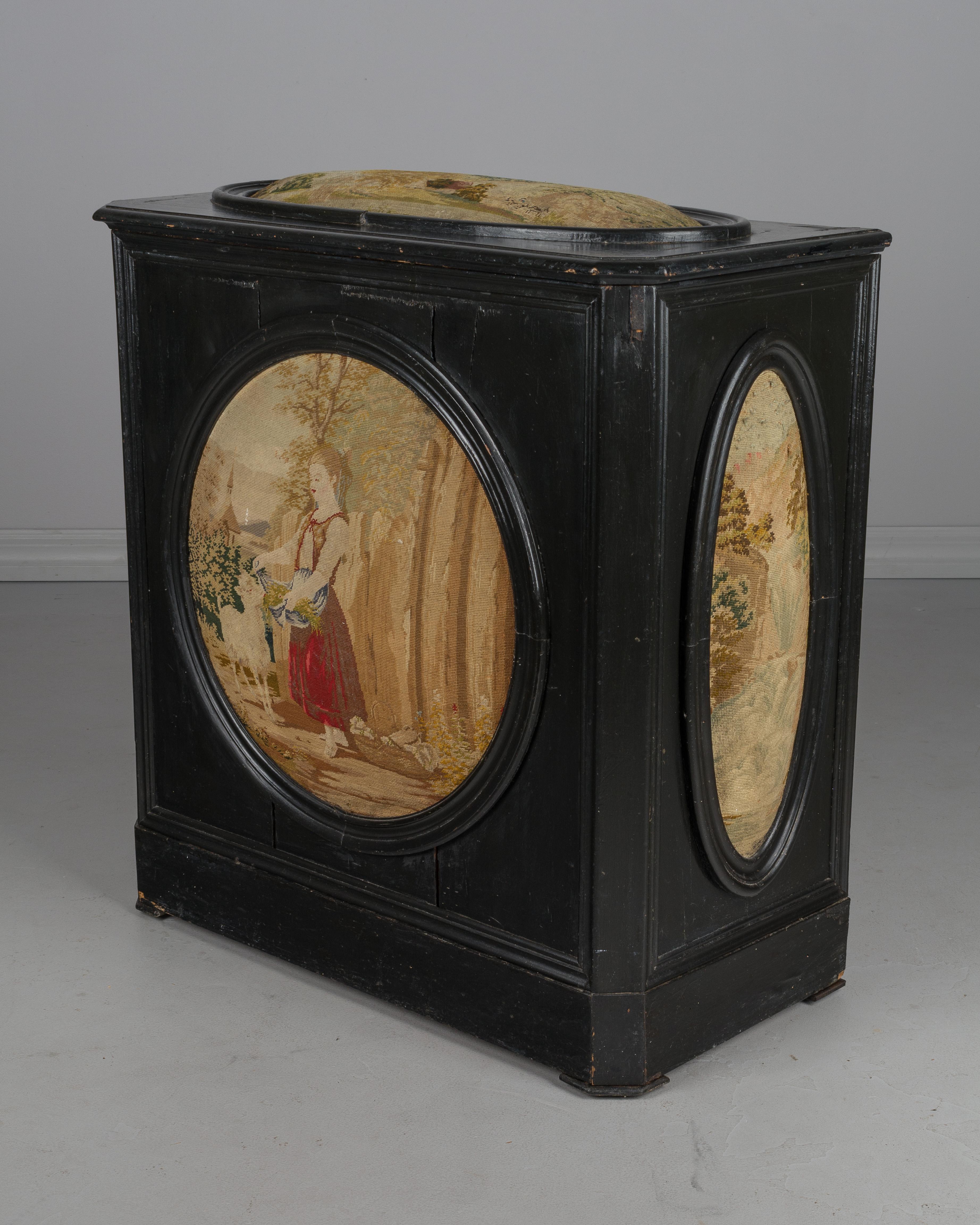 A 19th century French coffer, or storage hamper with upholstered petit point tapestry panels. Black painted pine with hinged top lid. Central image of a young maiden with lamb, side panels depicting waterfalls and the top with a mountain landscape.
