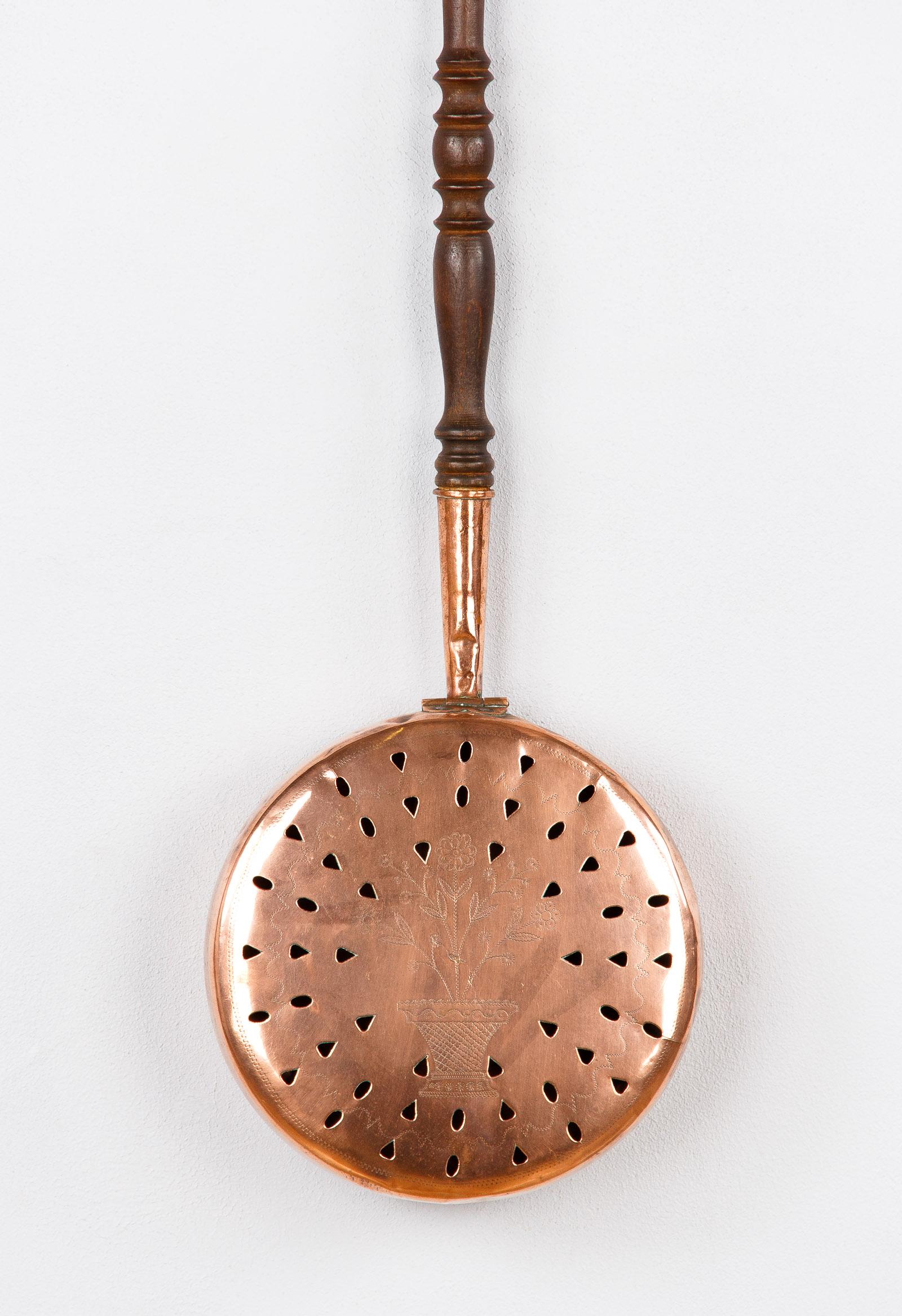 A copper bed warmer with a wooden handle from the Bresse region. The copper pan has a perforated lid and a sculpted motif of flowers in a pot.