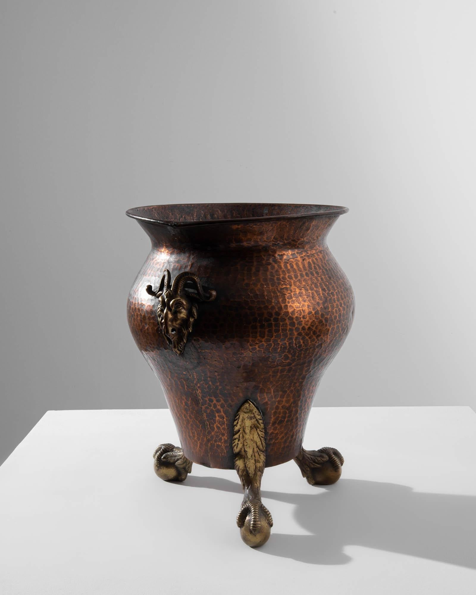 This antique copper vase offers a unique and intriguing accent. Made in France in the 1800s, the urn of beaten copper rests atop a trio of taloned brass feet; an intricately cast goat’s head in the center of the vase provides a compelling focal