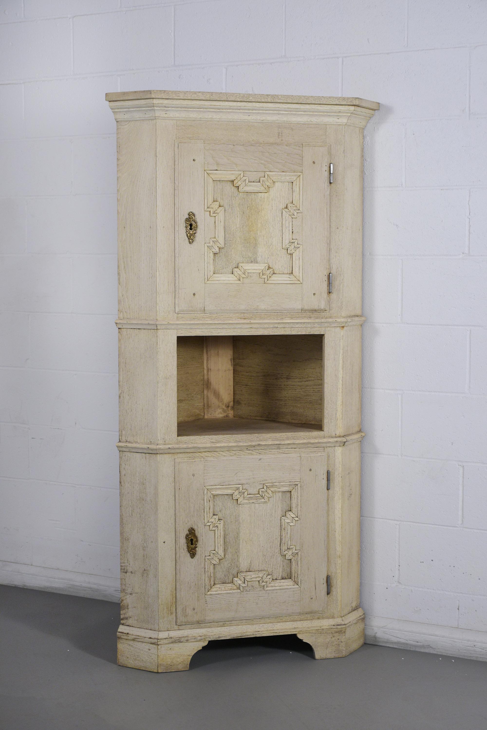 This antique French corner cupboard is made out of oak wood, has been given a newly bleached color finish, and is professionally restored. The corner cabinet has two doors with a carved panel design, original brass keyplates, and an open shelf in