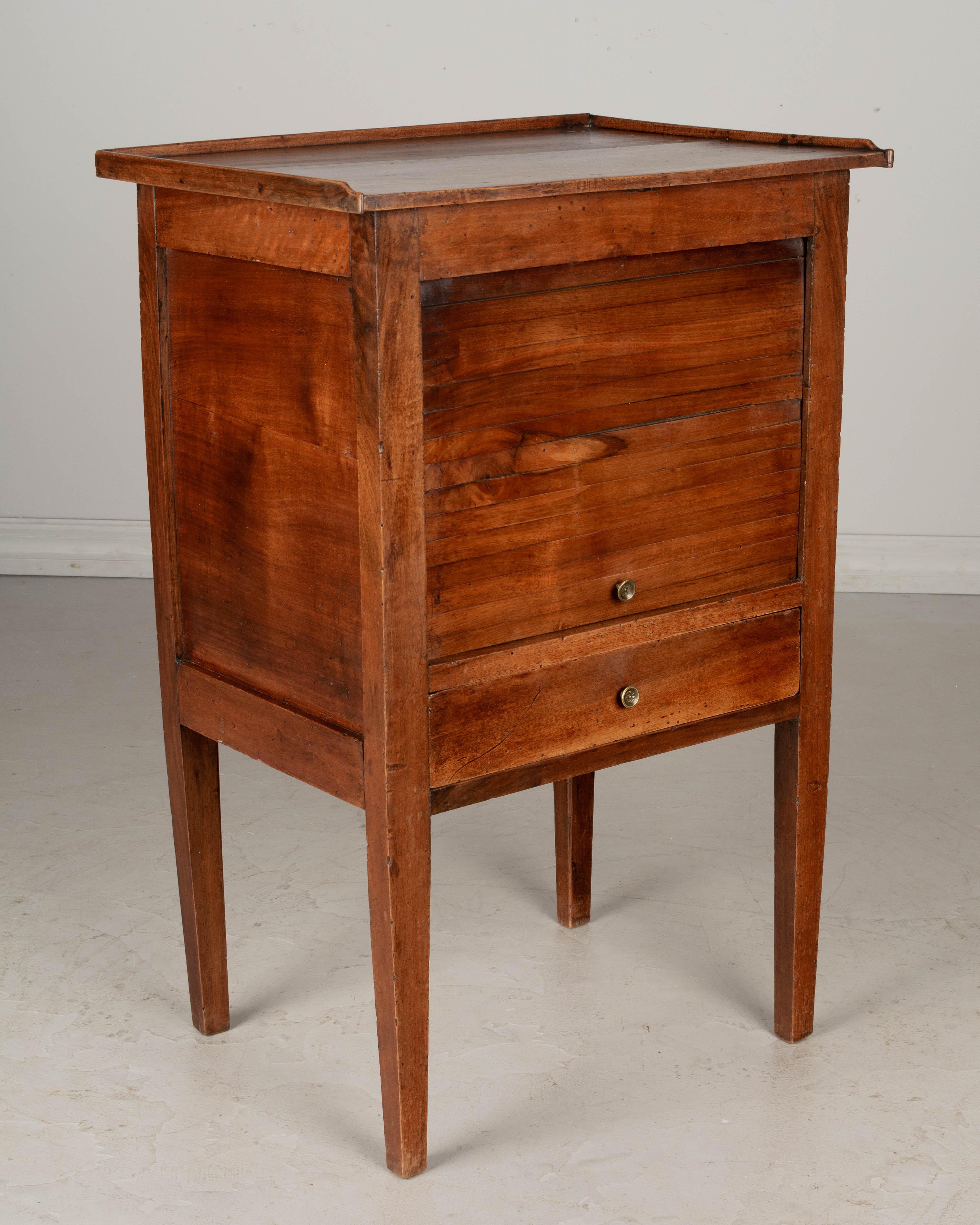 A 19th century French Country walnut side table with a vertical tambour door above a dovetailed drawer. Finished on all four sides. Small brass knobs. One interior shelf. Waxed patina. All original. May be used as a bedside table.