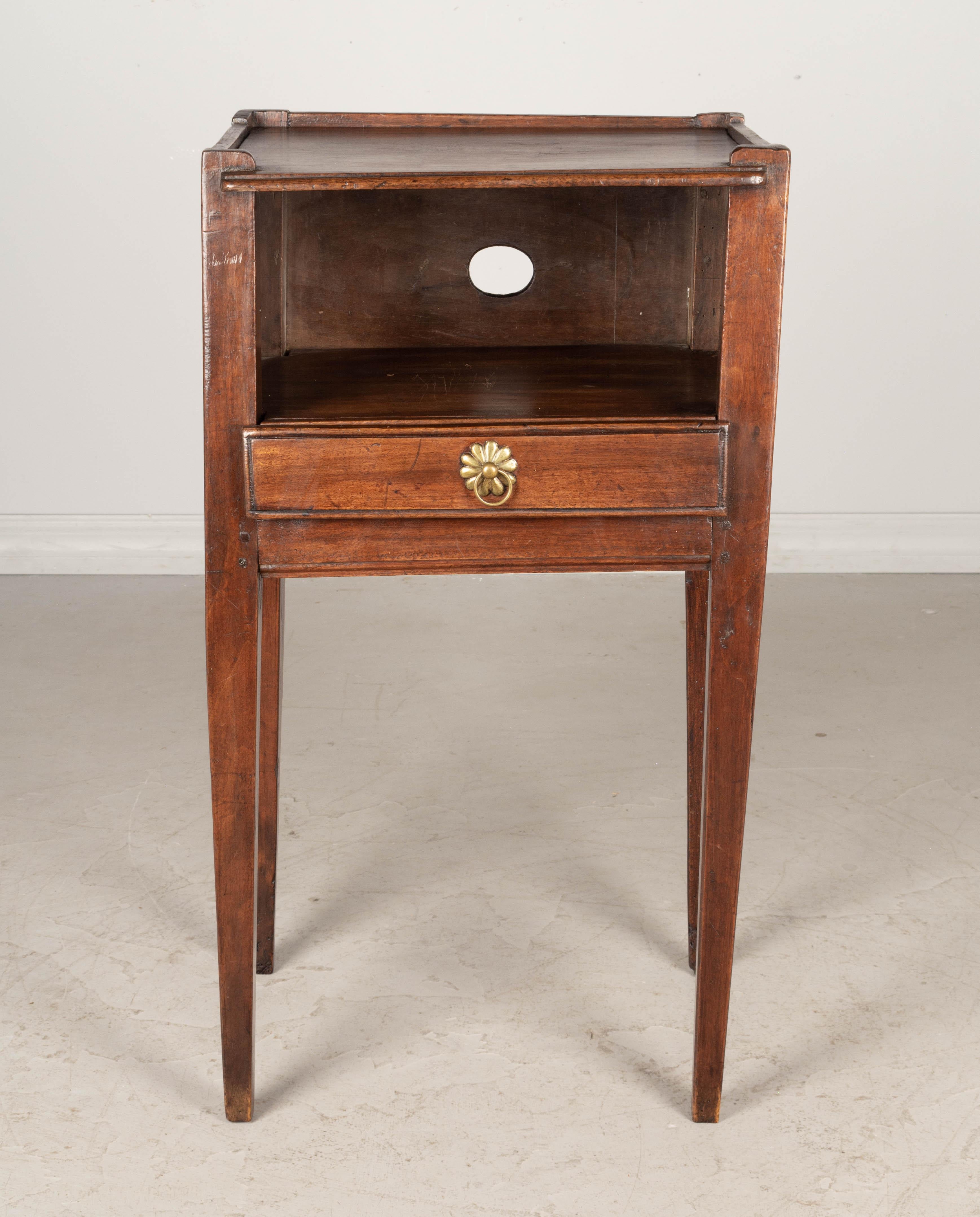 A 19th century Country French side table or nightstand made of solid walnut, with open niche and pierced quatrefoil shaped cut-outs on the sides. Drawer with brass ring pull. Slender tapered legs. Waxed patina.