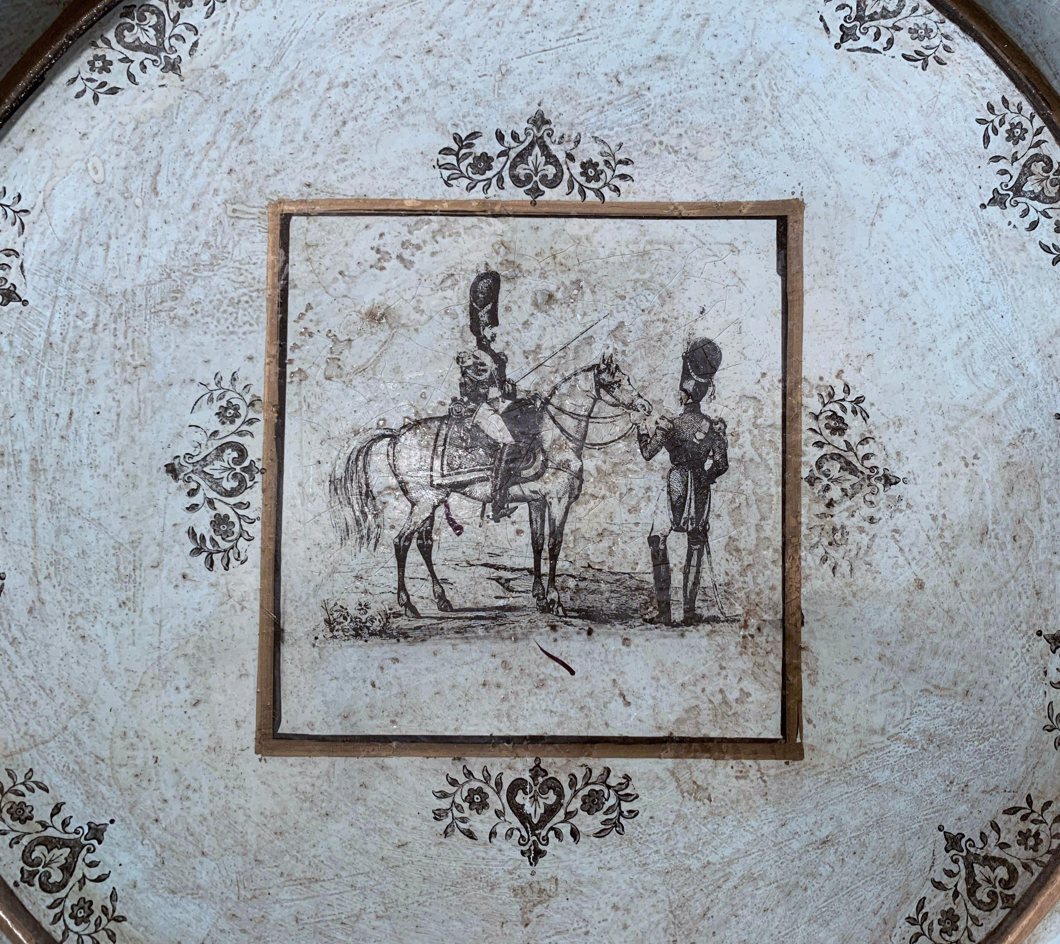 This elegant decorative tray was crafted in France, circa 1820, round in shape, the tole piece has hand painted decor with a center medallion featuring riders and horse in Directoire uniform; the decor is embellished with floral motifs around the