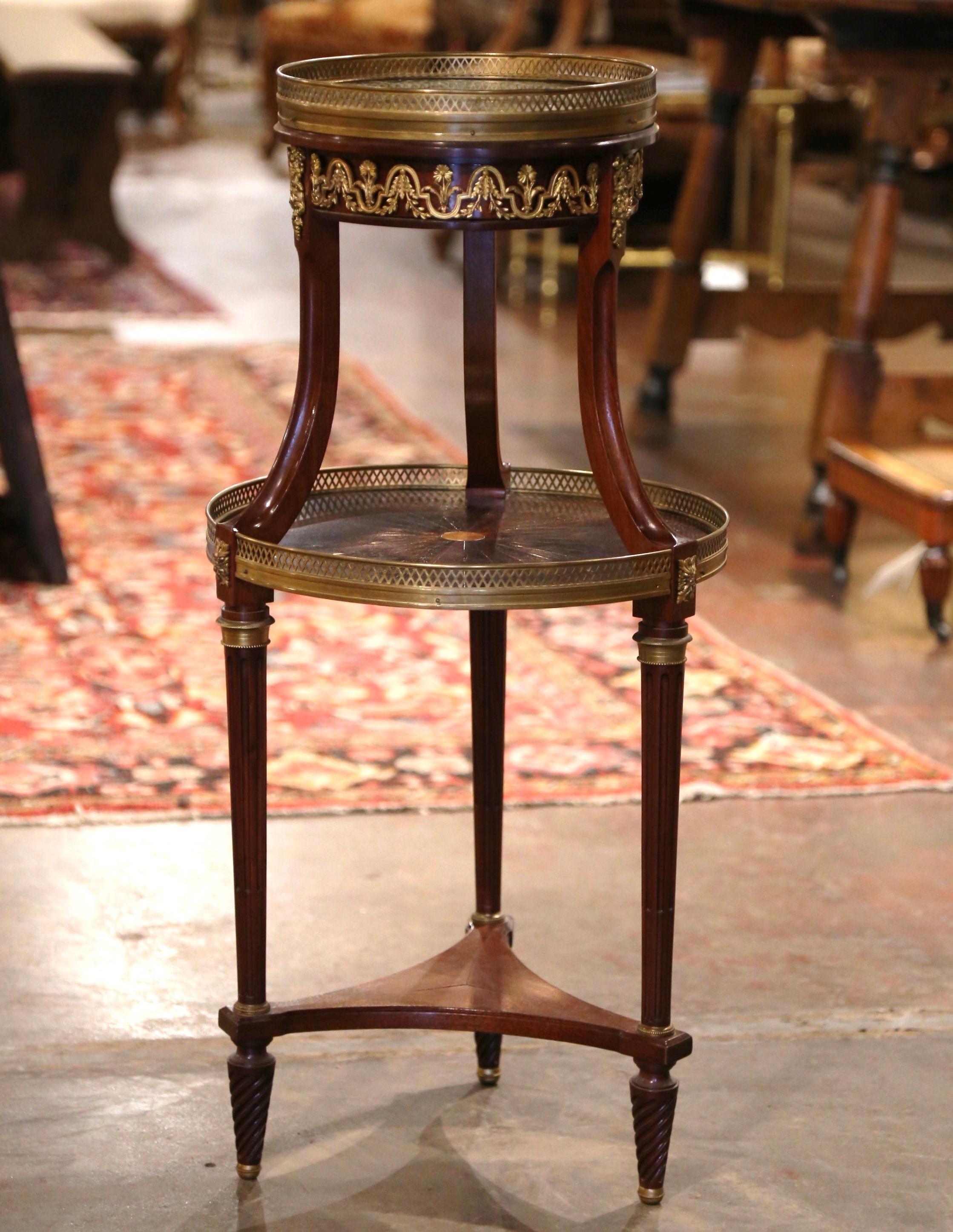 This petite and tall antique two-tier table was crafted in France circa 1880. Standing on three tapered and fluted legs decorated with brass rings and ending twisted feet over a triangle form stretcher, the round bouillotte table is decorated with