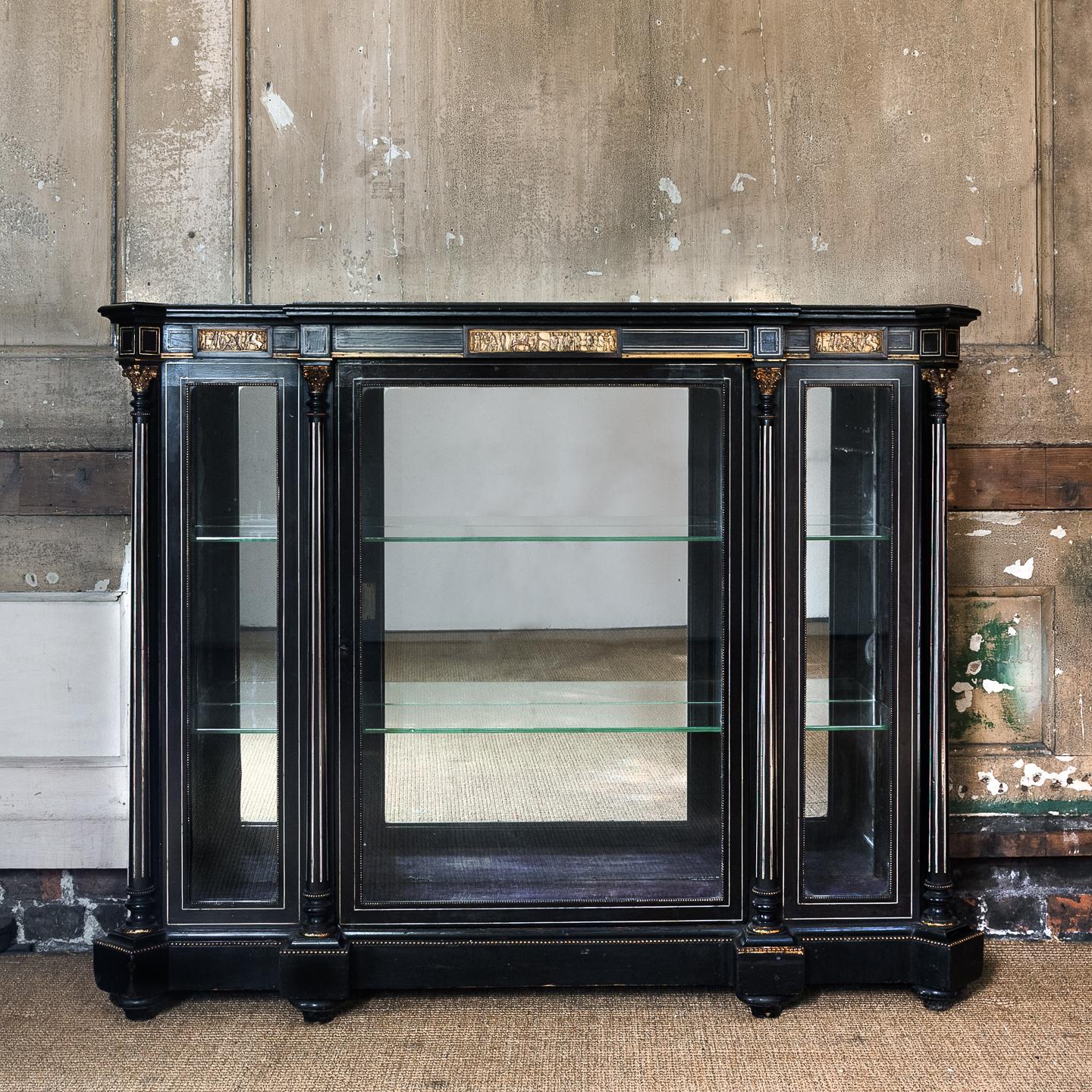 A mid-19th century French ebonized and inlayed display cabinet, of breakfront form, the (later) mirrored top above frieze with cast bronze Grecian bas-relief panels, the body with mirrored back and all-round glazing intersected by Corinthian