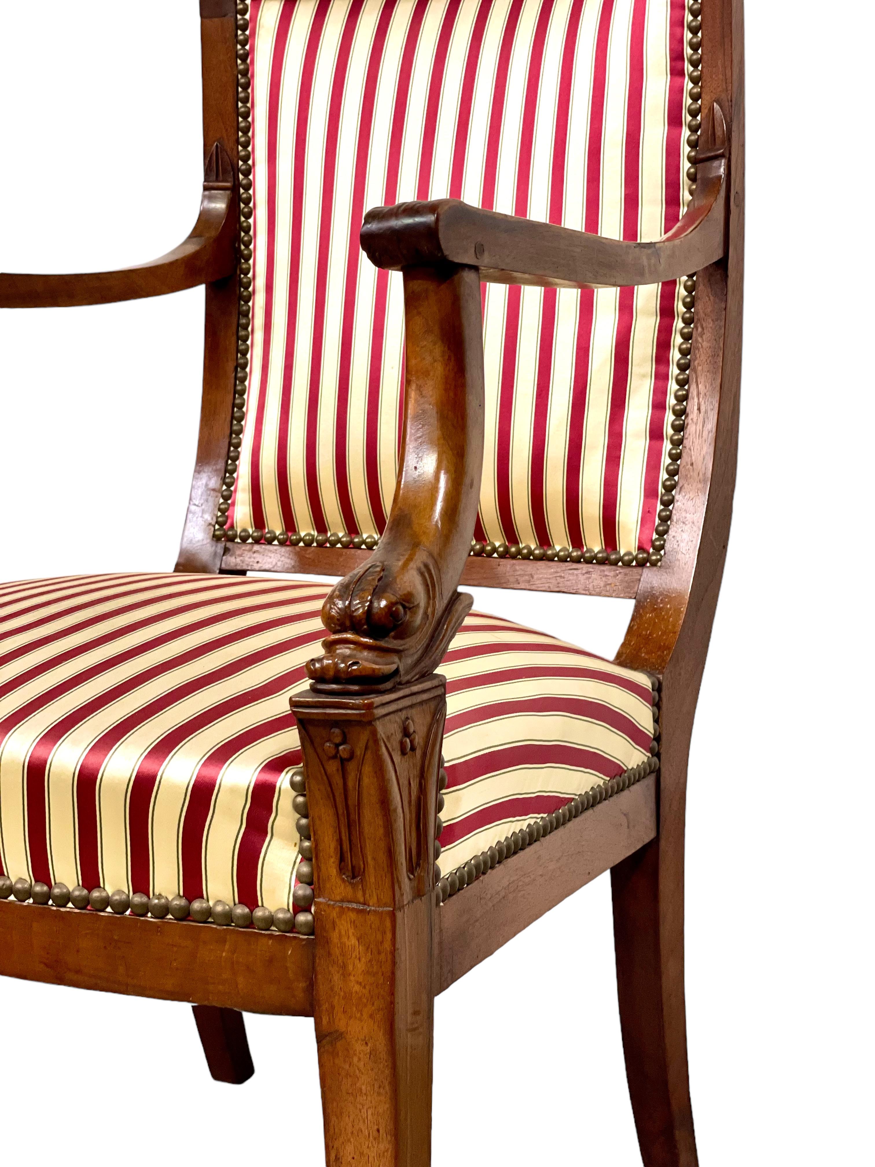 A fine Empire armchair with four elegant saber legs, upholstered in vertical cream and crimson stripes, and featuring ornately carved dolphins' heads at the base of each shaped arm rest. This stately and very handsome chair has a wide, comfortably