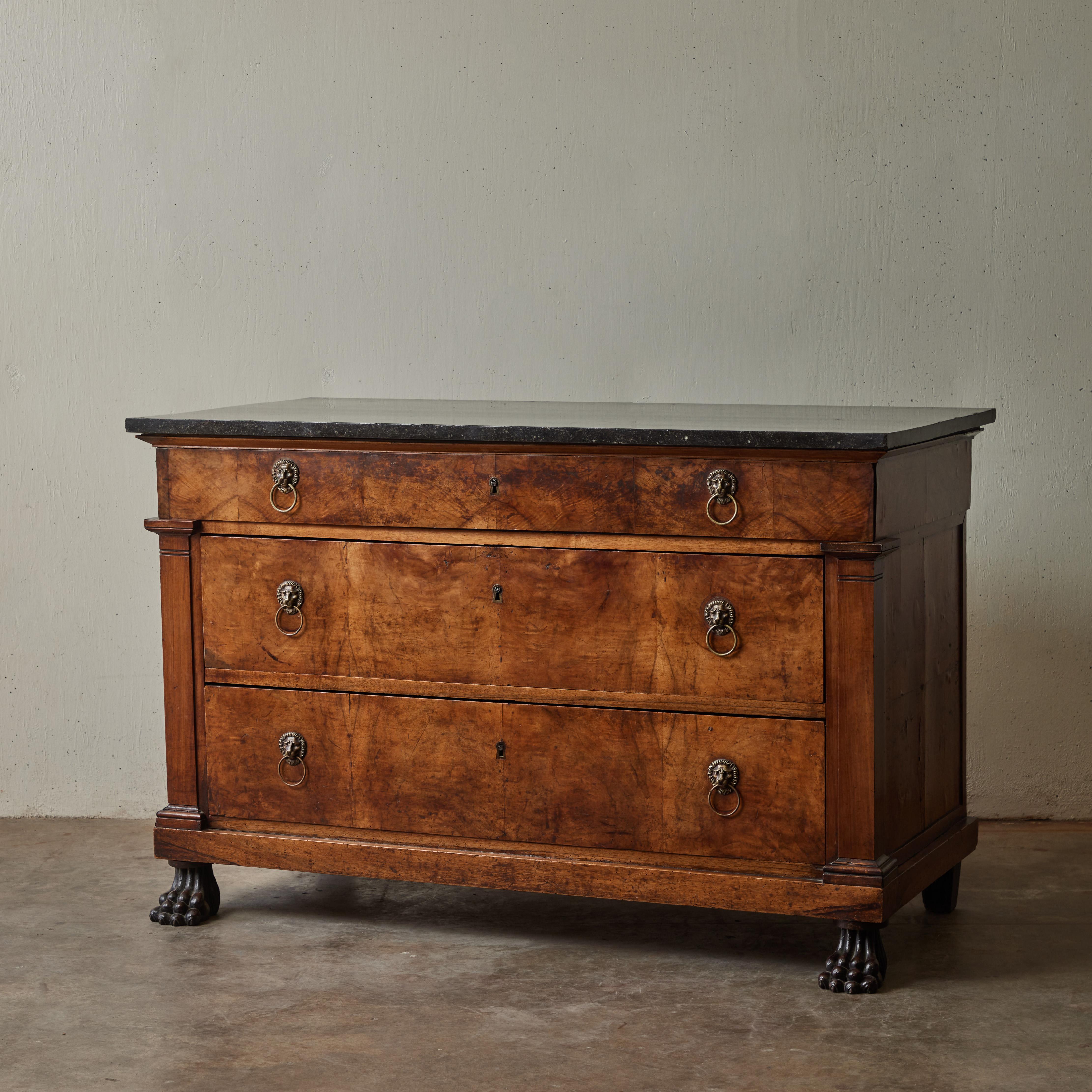 An early 19th-century French Empire style chest of drawers in walnut with black marble top, carved claw feet, and brass lion's head ring-pull handles. The warm, honey-toned wood has a rich visual texture, which complements that of its top. The
