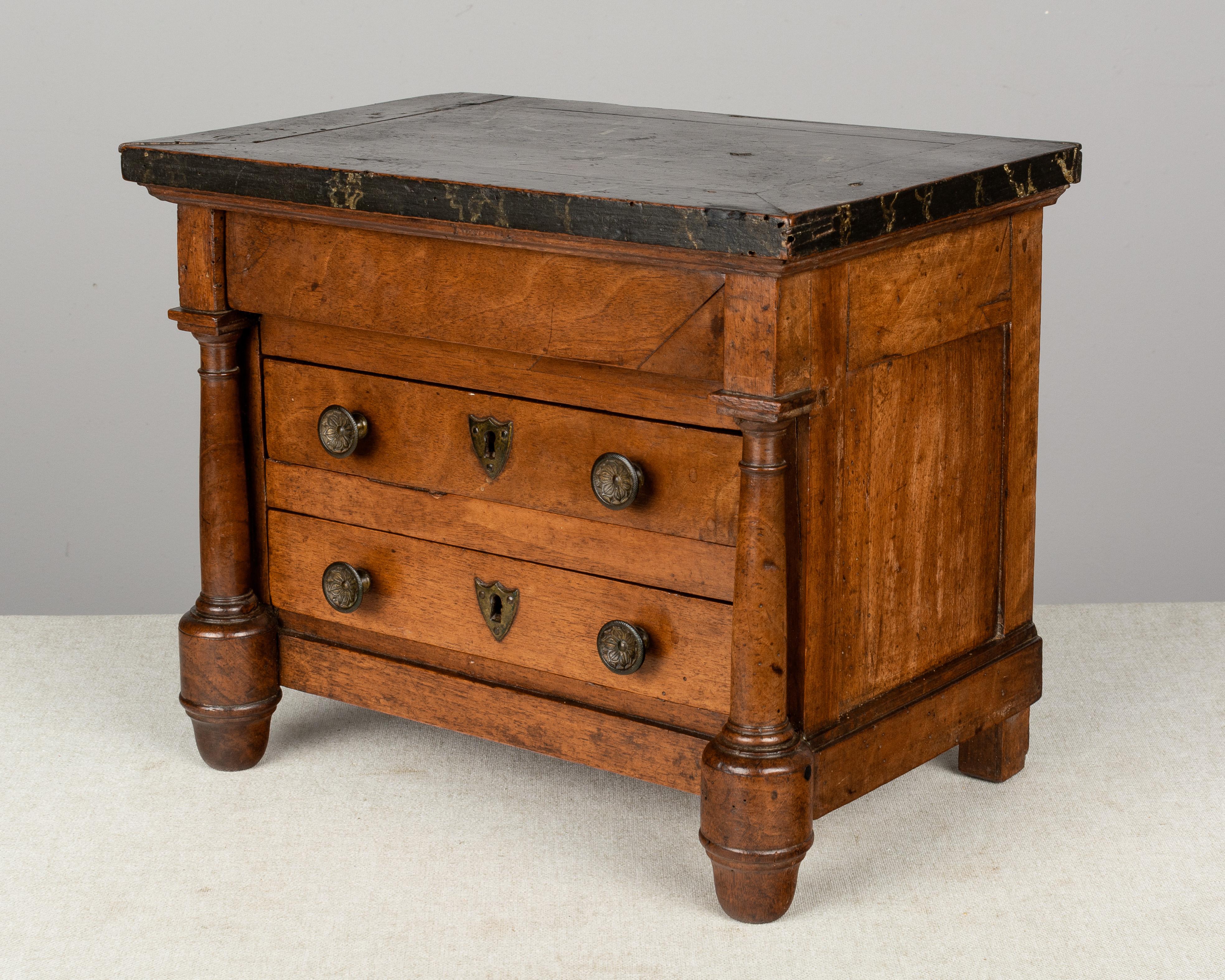 An early 19th century French Empire miniature sample commode made of solid walnut with three dovetailed drawers, turned columns and a top that is faux painted to look like veined black marble. Original brass knobs and escutcheons. Locks were never