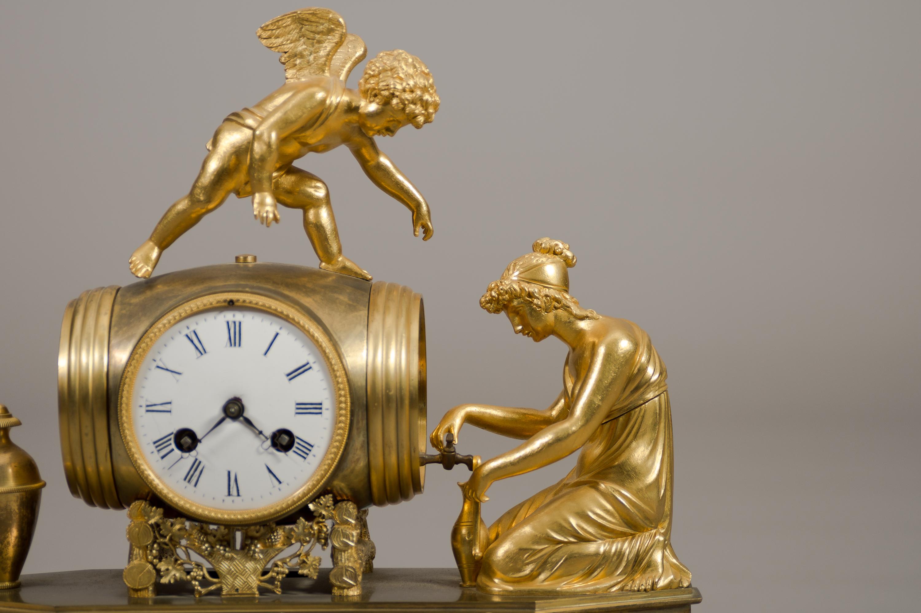 A fine Early 19th century French Empire ormolu bronze mantel clock. 

Depicting a winged cherub standing atop a barrel while a kneeling lady pouring wine into a vase. Having a white enamel dial with Roman numerals, and a movement with a silk wire