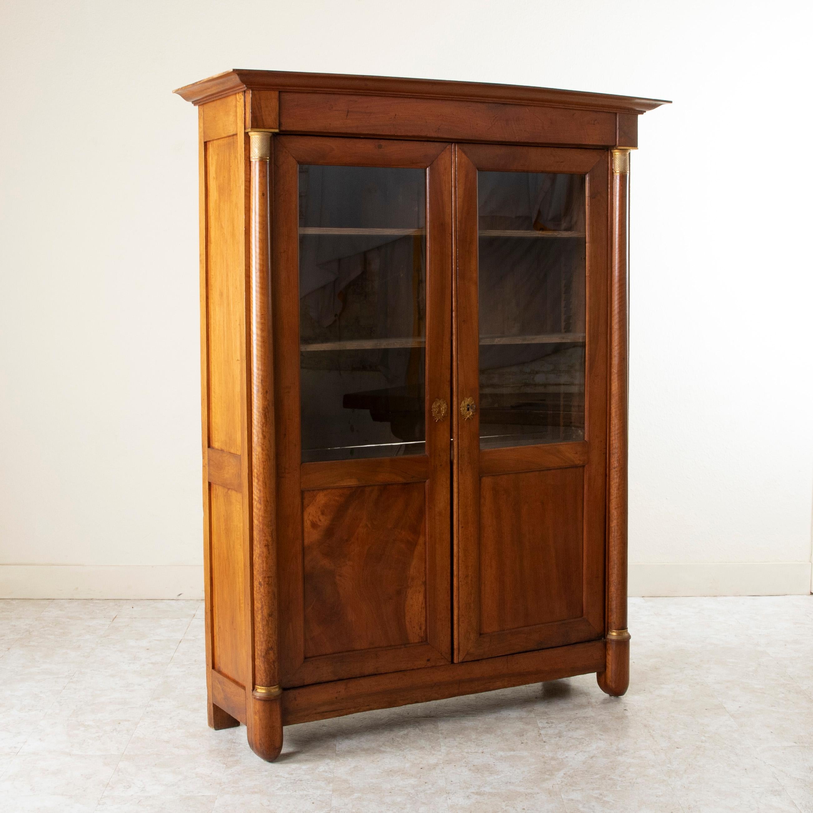 This early nineteenth century Empire period walnut bookcase features half columns that flank its glass doors. The columns are capped in bronze detailed with diamond and a Vitruvian scroll pattern. The base of the columns are additionally detailed in
