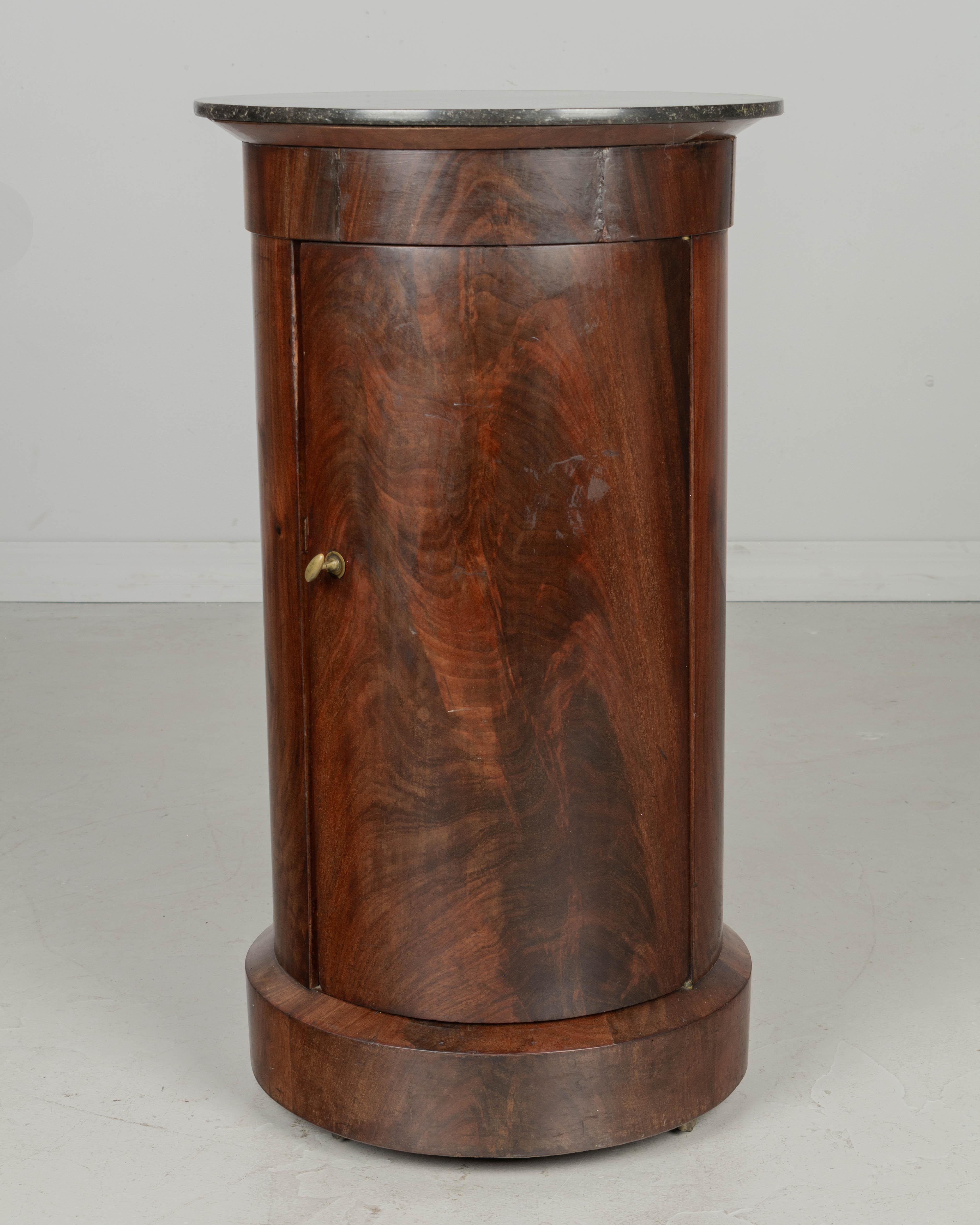 A 19th Century French Empire style circular side table, or nightstand, made of beautifully patterned veneer of mahogany on pine with a dark gray St. Anne marble top. Door with small brass knob opens to two interior shelves. Four brass wheels, rolls