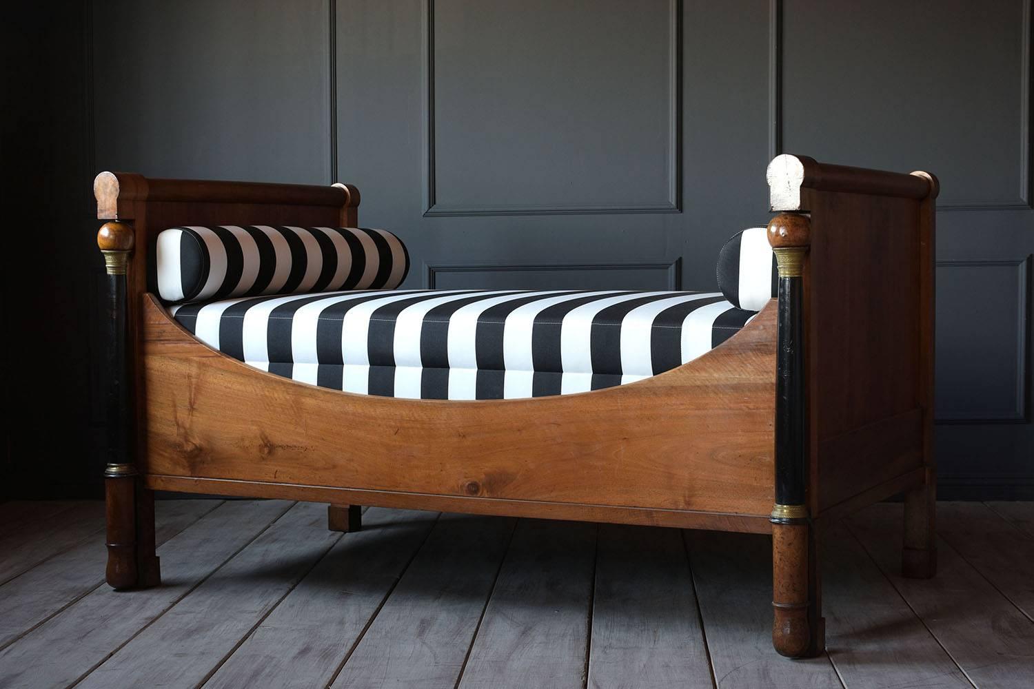 This 1840s French Empire-style daybed features a walnut wood frame with a black and walnut stained finish. The rectangular head-board and foot-board have a rounded upper bar and carved columns on the side with bronze ornaments on the top and bottom.