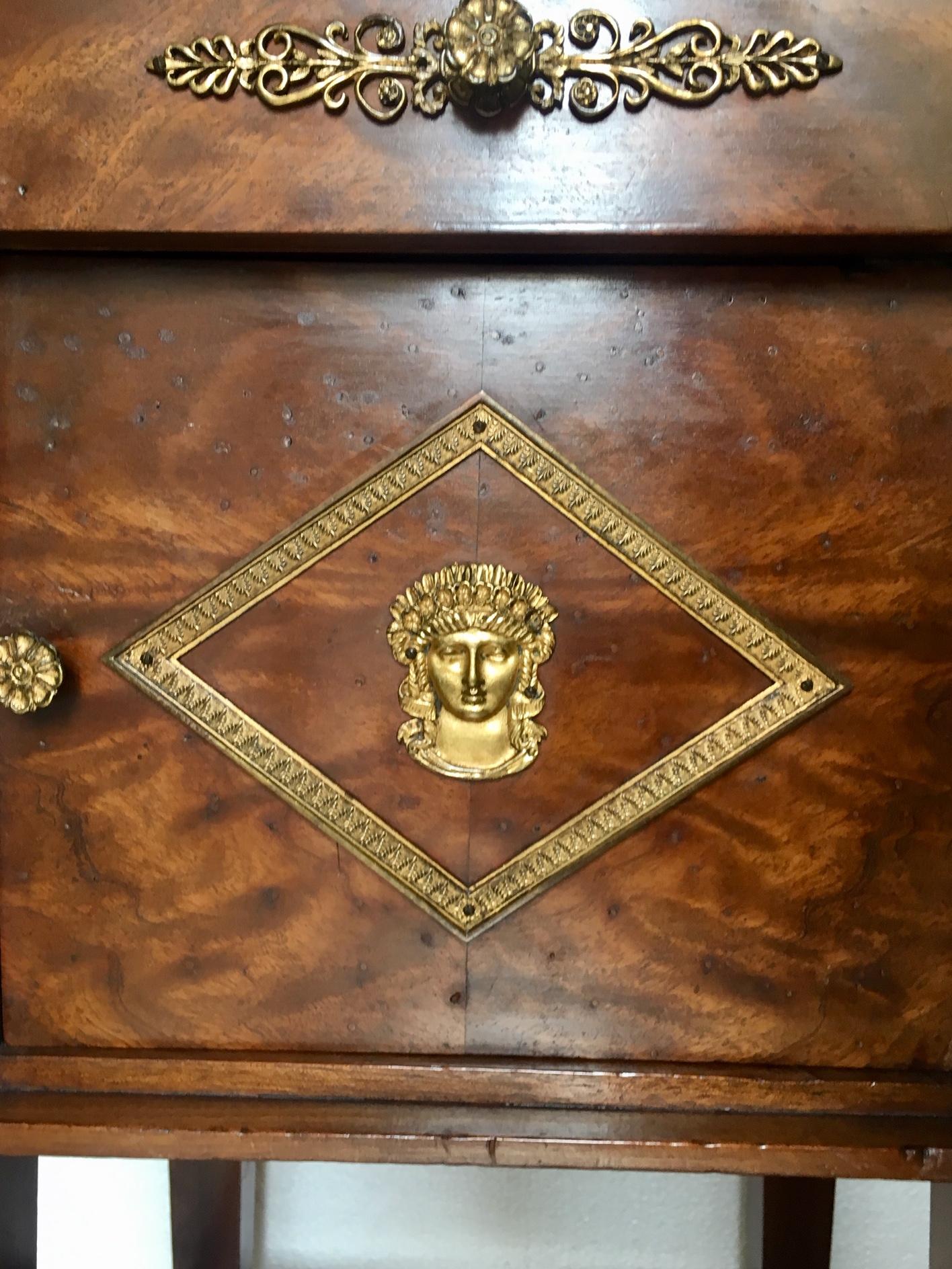 French Empire side or night table, in walnut wood and original black marble top, a drawer and a cabinet door, with original classic gilded bronze, representing leaves, capitals on its edges, and a female face in the center of the door, legs that end