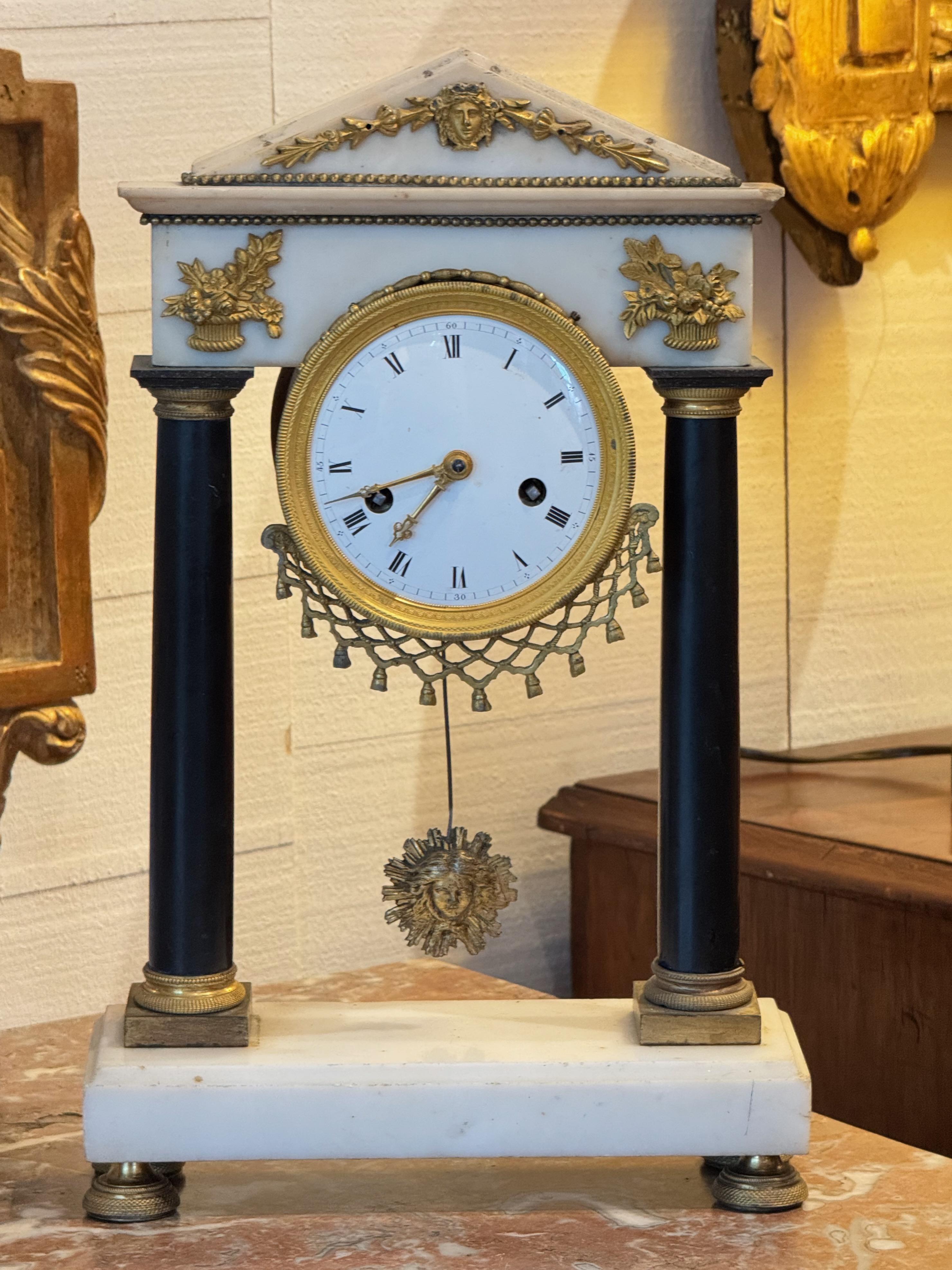 Orologio da camino francese in stile Impero, caratterizzato da una cassa in marmo bianco con supporti a colonna neri e classici accenti in bronzo dorato (ormolu). Il frontone triangolare è decorato con un mascherone centrale e con delle volute di