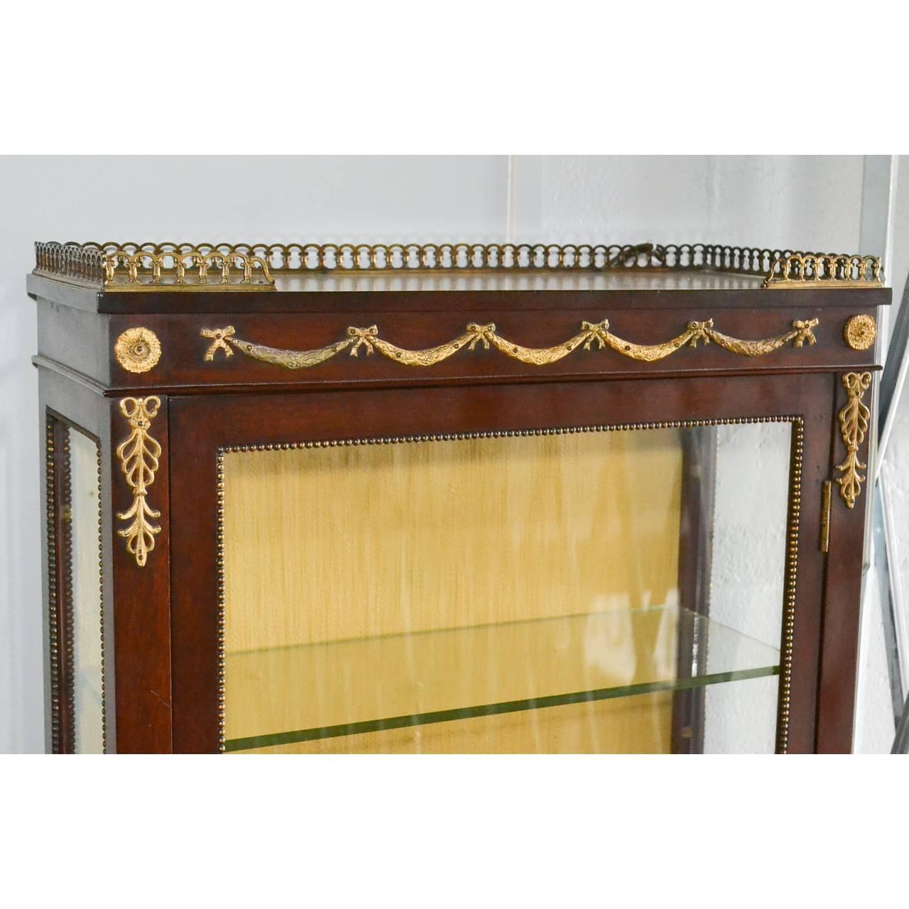 Stunning 19th century French Empire design mahogany vitrine cabinet with a partial brass gallery above a frieze with exquisite gilt bronze ribbon and swags. The bronze trimmed glass door rests on a base with a fitted drawer supported by ebonized and