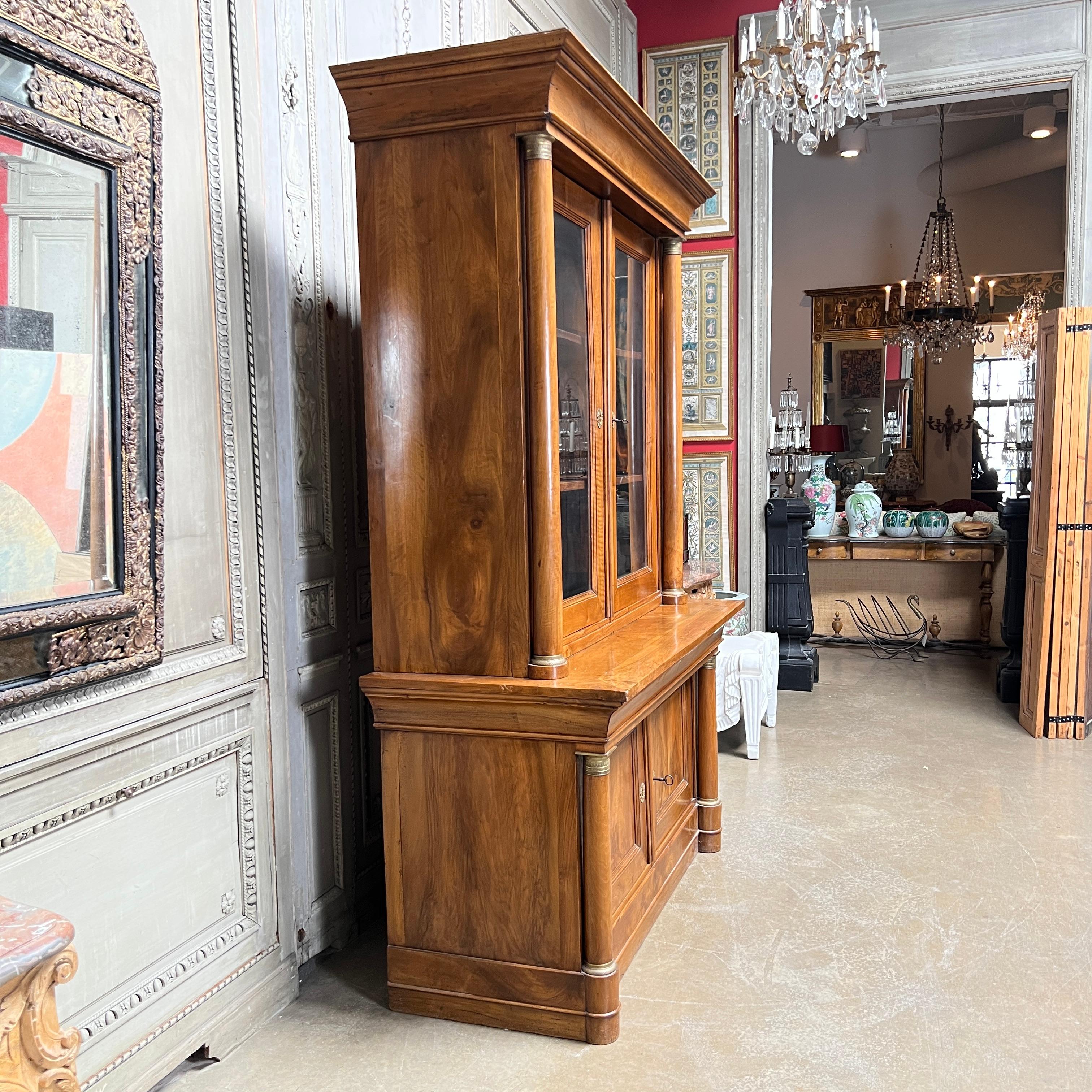 A large French Empire bookcase, or buffet a deux corps in walnut with bronze mounts and glass doors, dating from the early 19th century. 
This handsome piece has a nice old patina and well cast bronze mounts. It would make an ideal bookcase, house