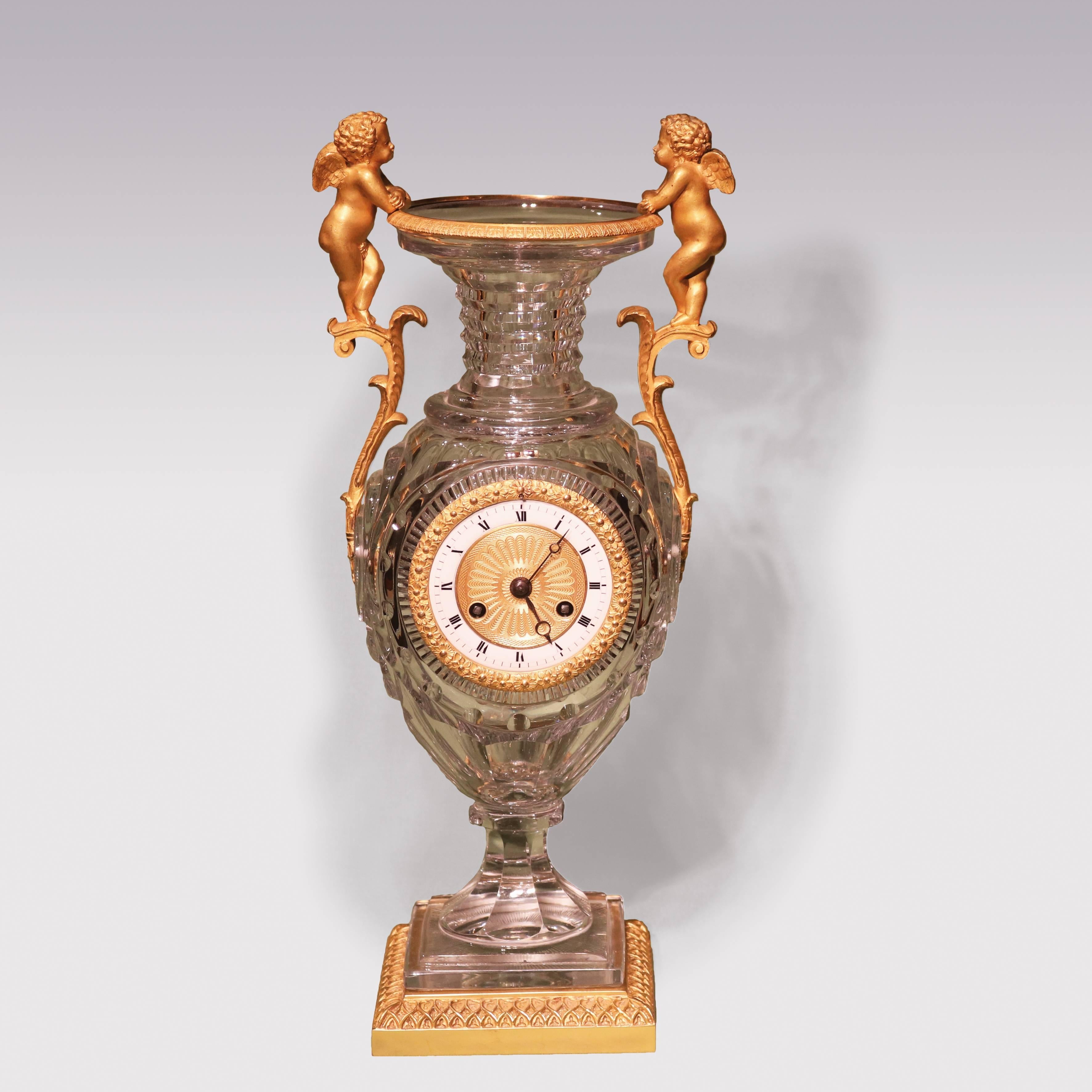 An early 19th century French Clock, having enameled dial 8-day striking movement, contained in unusual faceted glass urn with ormolu rim and cherub scroll handles, ending on square plinth base with ormolu border.