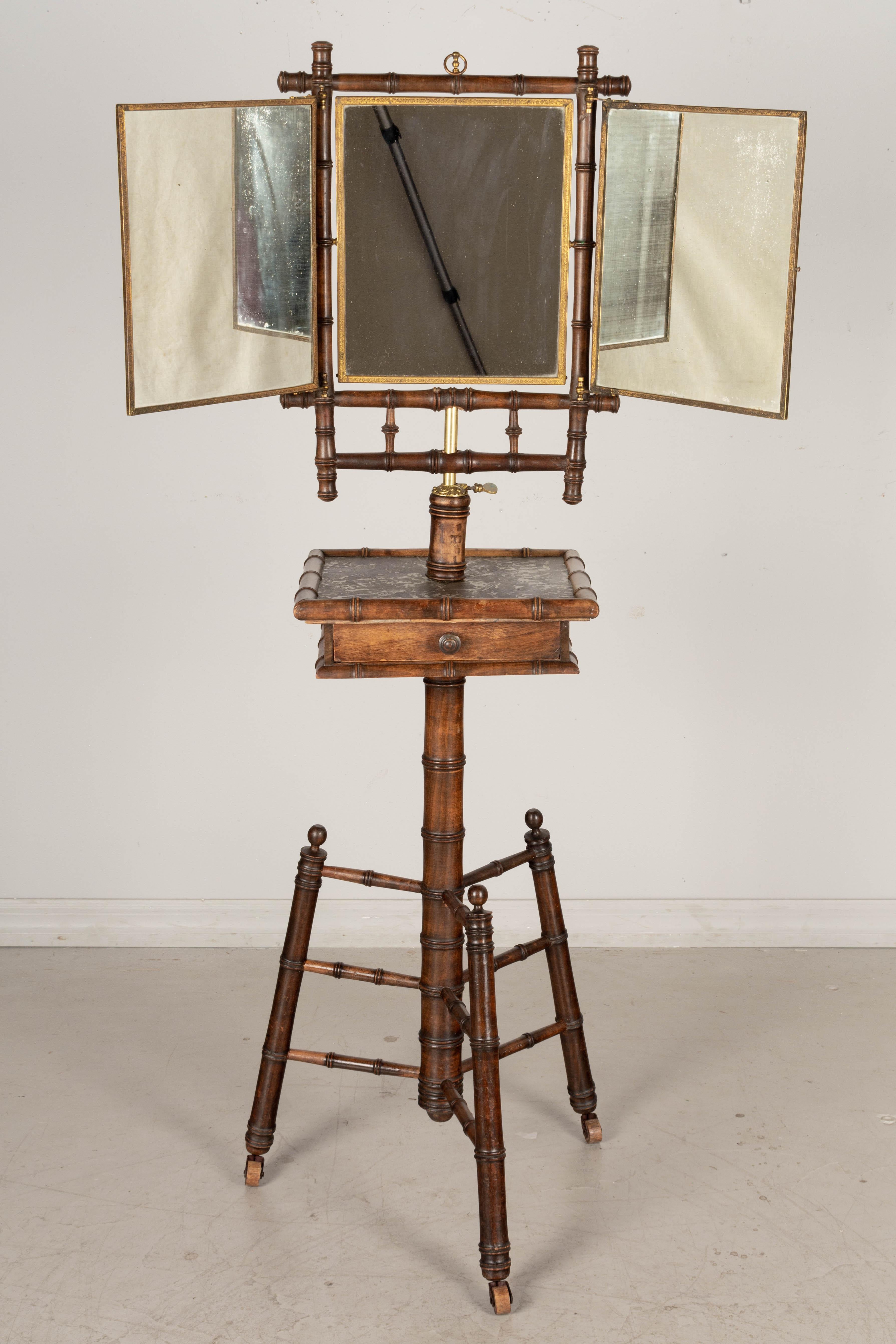 A 19th Century French faux bamboo cherry wood stand with folding mirror. Sturdy tripod base with divided drawer and marble top. Trifold mirror has brass trim and is backed with decorative anaglypta, a heavy paper with an embossed floral motif.