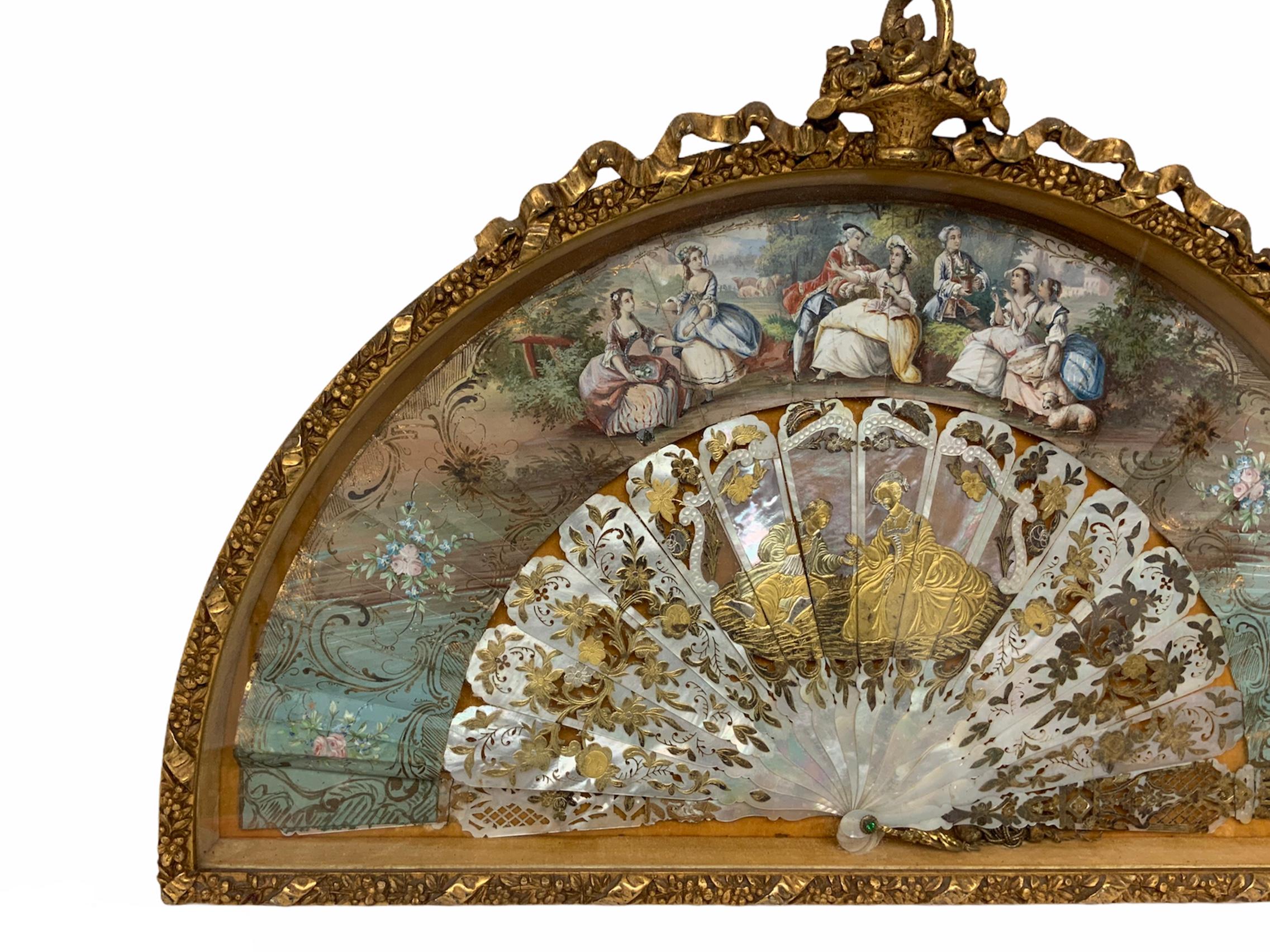 This is a hand painted ladies fan with an 18th century scene of a courting couple in a pastoral landscape. Also the reticulated mother of pearls sticks depict a gilded scene of a courting couple surrounded by flowers. The fan is framed in a gilded