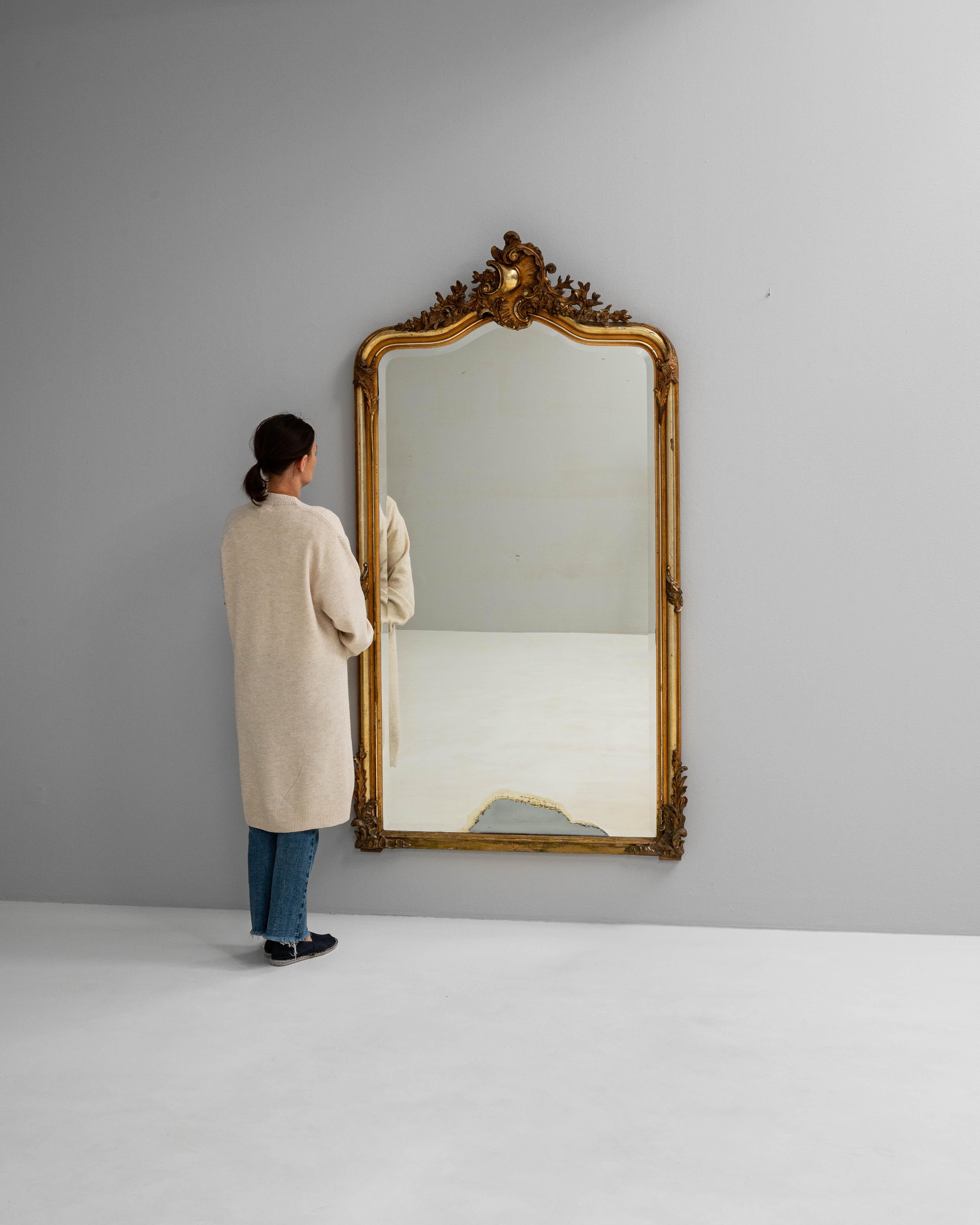 Este espejo francés del siglo XIX presenta un marco de madera dorada elaboradamente tallado, coronado con una cresta ornamentada de follaje en volutas y motivos florales de estilo rococó. El acabado dorado ha envejecido con gracia, mostrando una