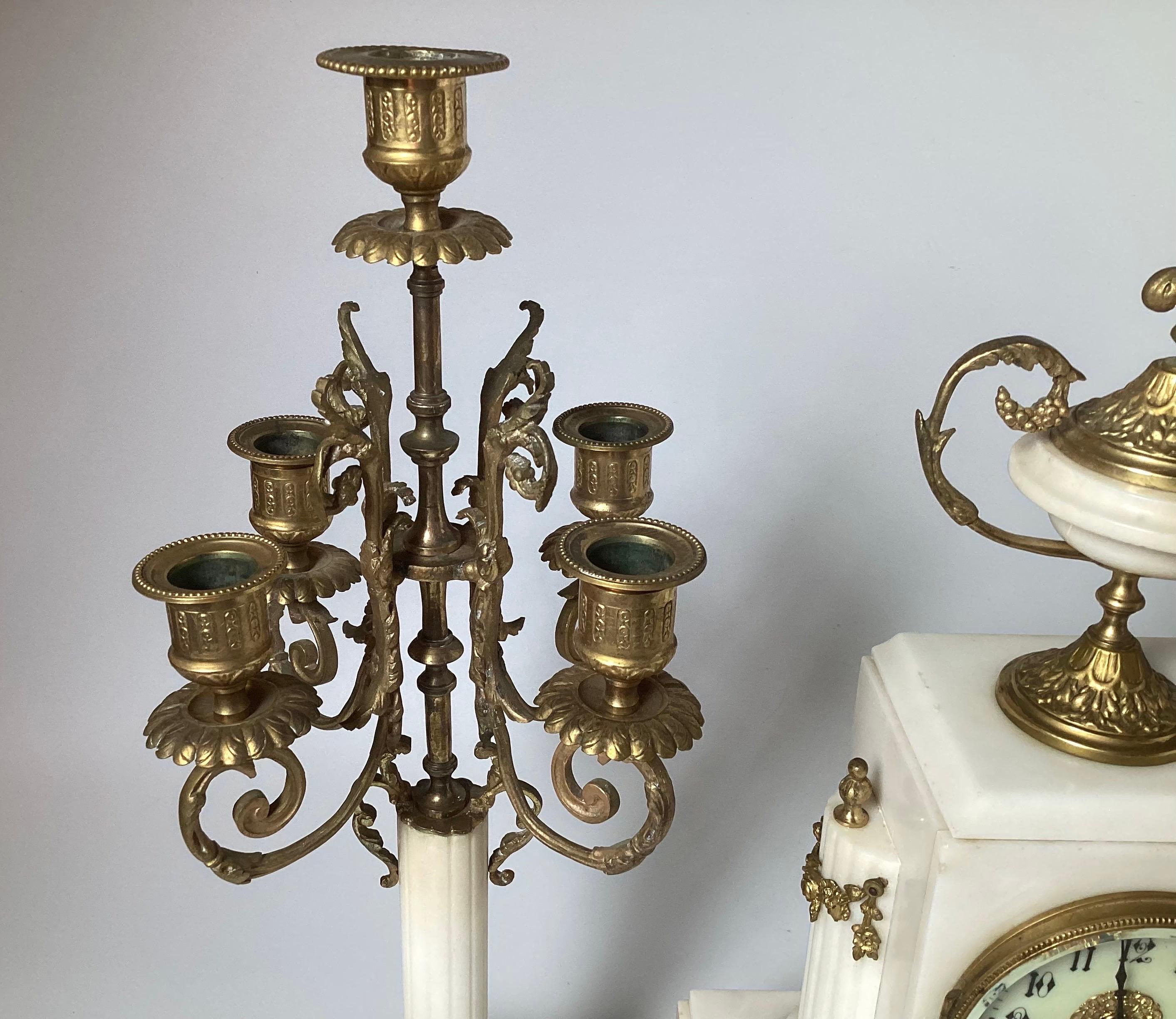 Orologio francese in bronzo dorato e marmo bianco in tre pezzi, fine del XIX secolo. Comprende un orologio da camino e una coppia di candelabri a cinque luci. L'orologio ha un quadrante circolare smaltato bianco con numeri arabi, il movimento a