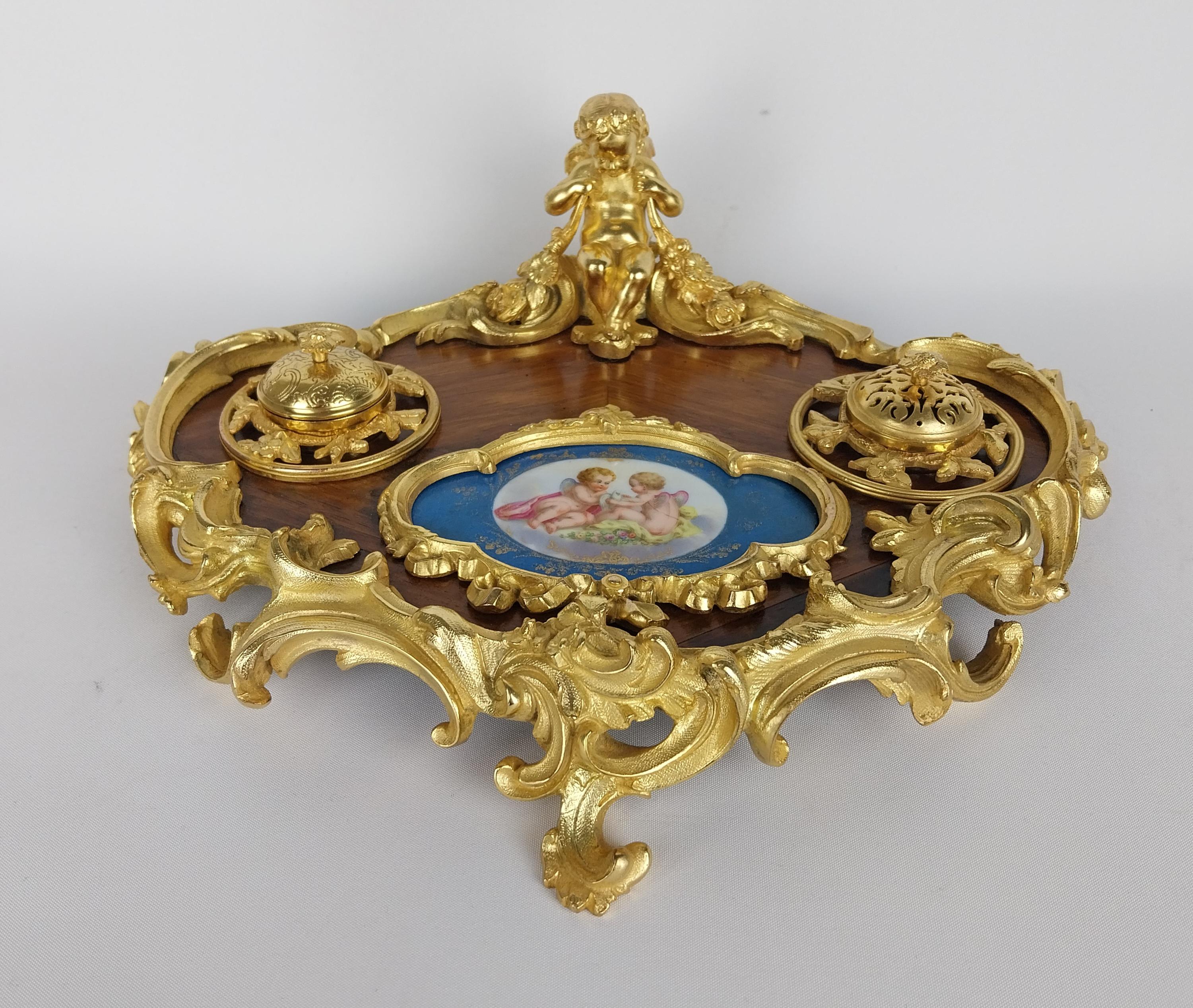 Supporto da scrivania in legno di tulipano francese con bordo in bronzo dorato in stile rococò,  un cherubino alato è seduto con in mano ghirlande floreali e guarda una placca in porcellana di Sèvres dipinta a mano che raffigura una coppia di