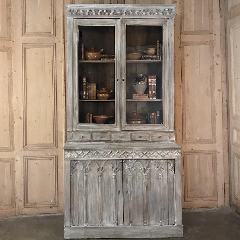 19th century French Gothic whitewashed bookcase features intricately carved trefoil and Moorish arch detailing, with spacious cabinets on the lower tier, and a tier of five drawers separating the upper glazed display case from the lower storage.