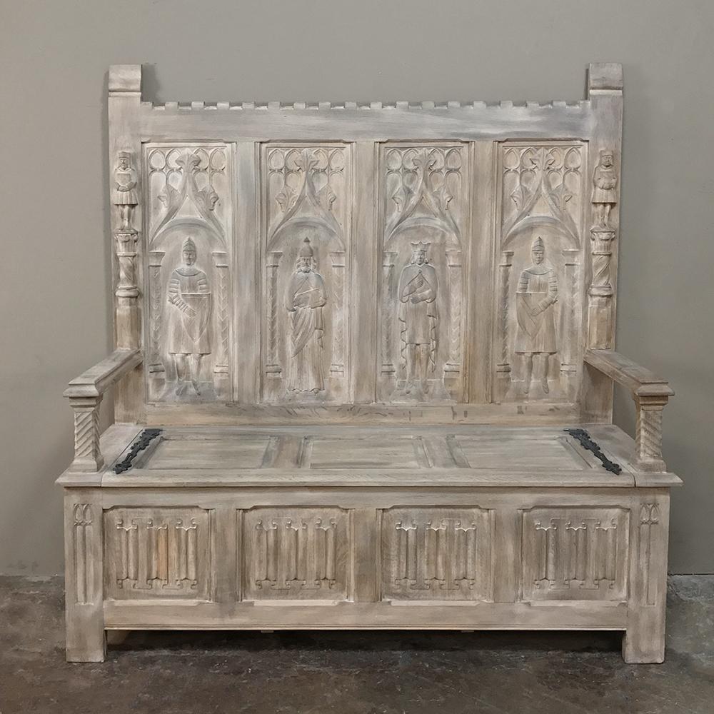 This timeless 19th century French Gothic whitewashed hall bench features four unique hand carved motifs on the seatback panels above which contrast with the consistent linenfold panels which appear on the trunk base below. Castellated seatback crown