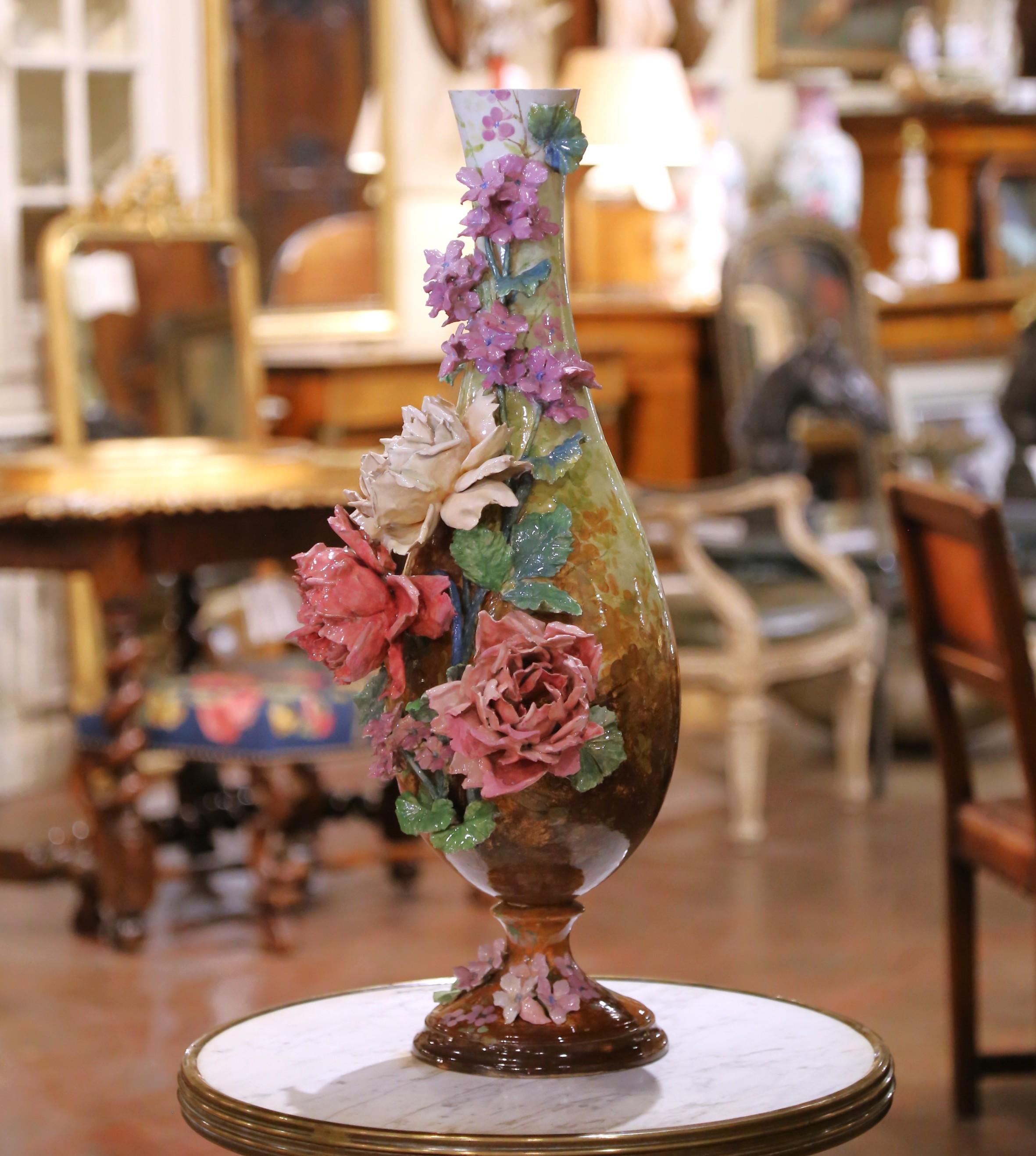 Questo impressionante vaso antico di Barbotine francese è un esempio magistrale di arte della faience, che si distingue per i suoi rilievi floreali riccamente applicati e la sua smaltatura pittorica. Alto 27 pollici e con un diametro di 11,5