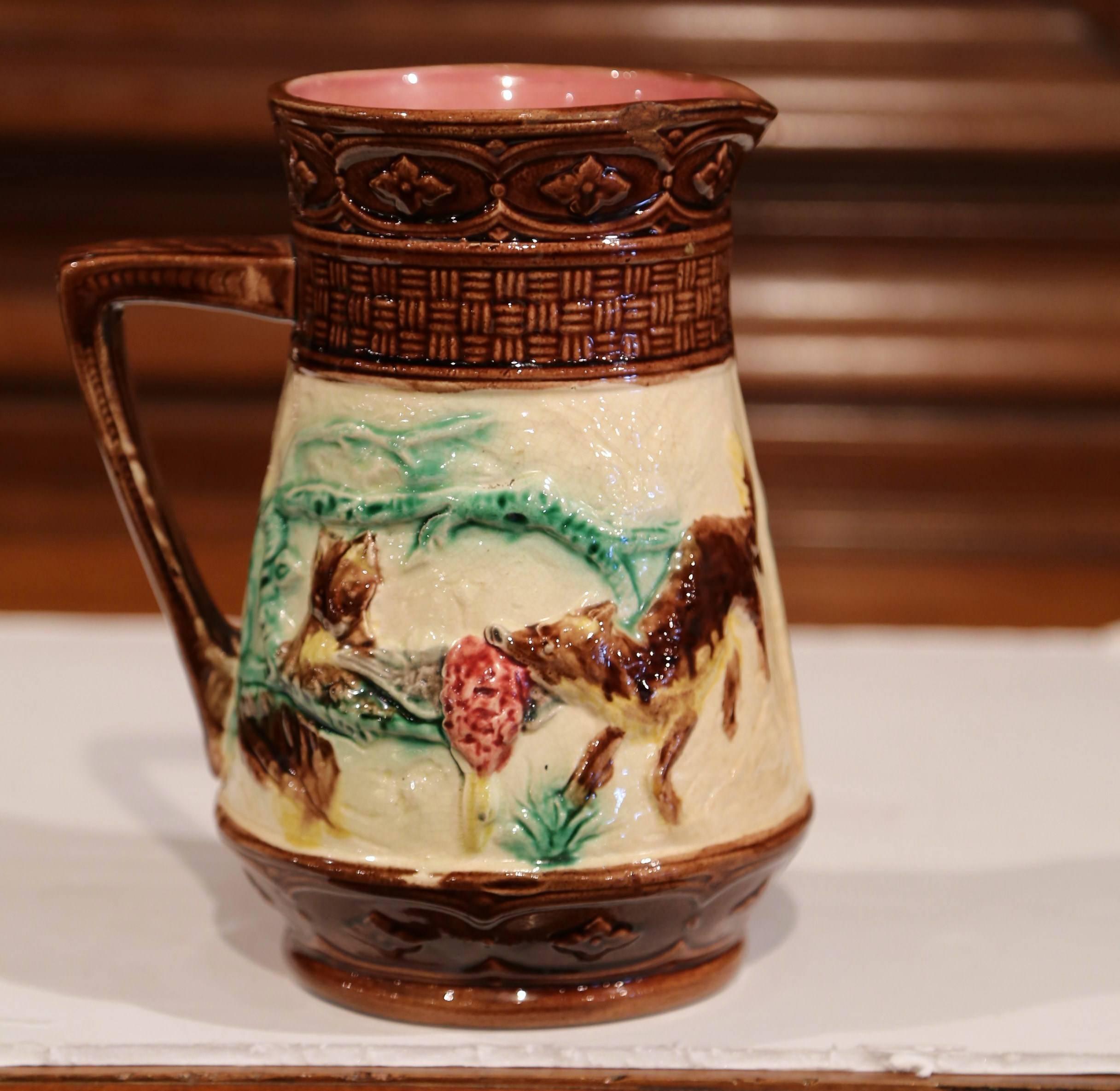 Crafted in France, circa 1880, this interesting antique pitcher would make a charming addition to your kitchen or dining room. The artful, hand painted, Majolica pitcher is embellished with a hunting scene, which shows a dog in high relief holding a