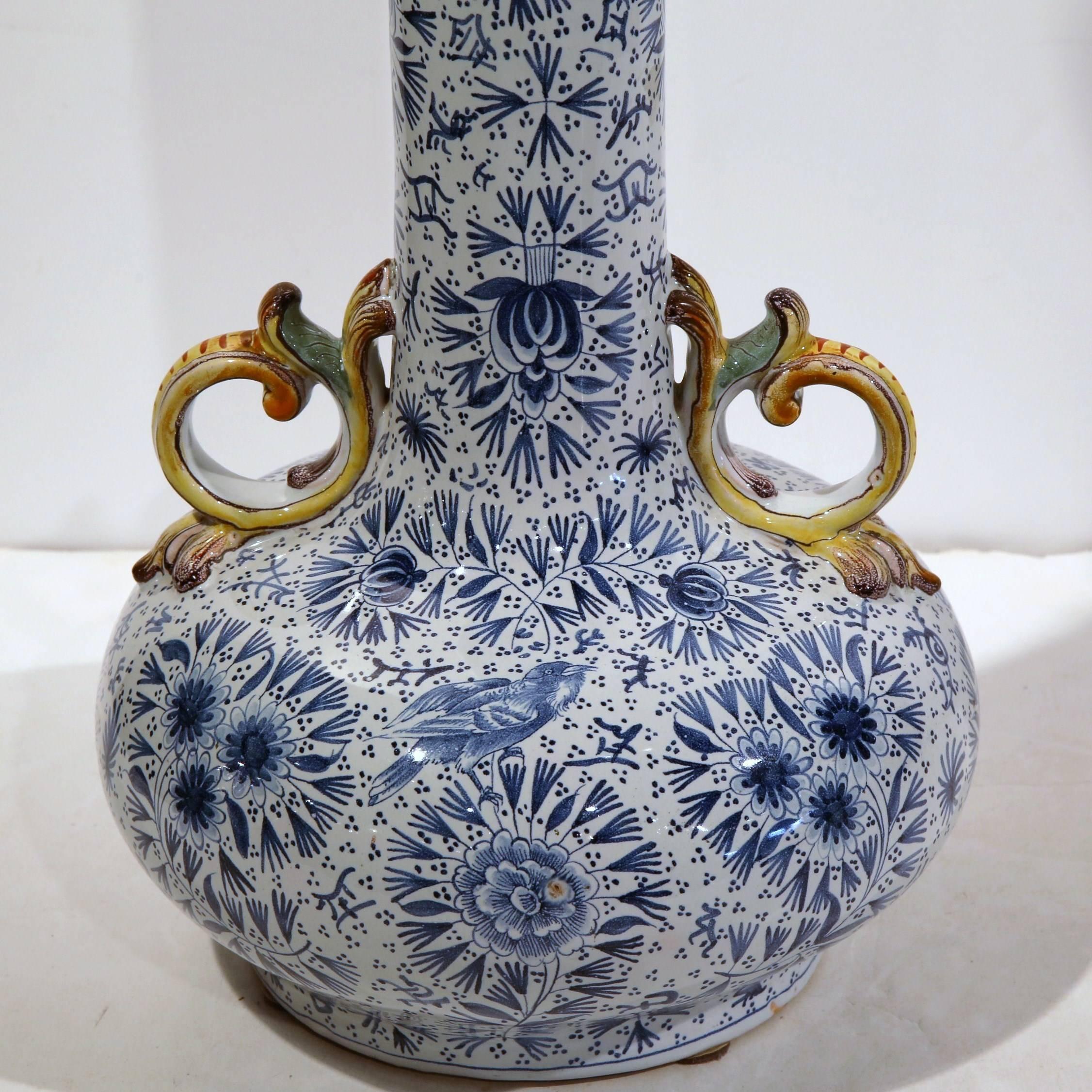 This elegant, antique vase was created in Luneville, France, circa 1870. The tall, thin vase has a Classic blue and white palette and is decorated with natural motifs including birds and flowers. The long neck is flanked by yellow dolphin handles at