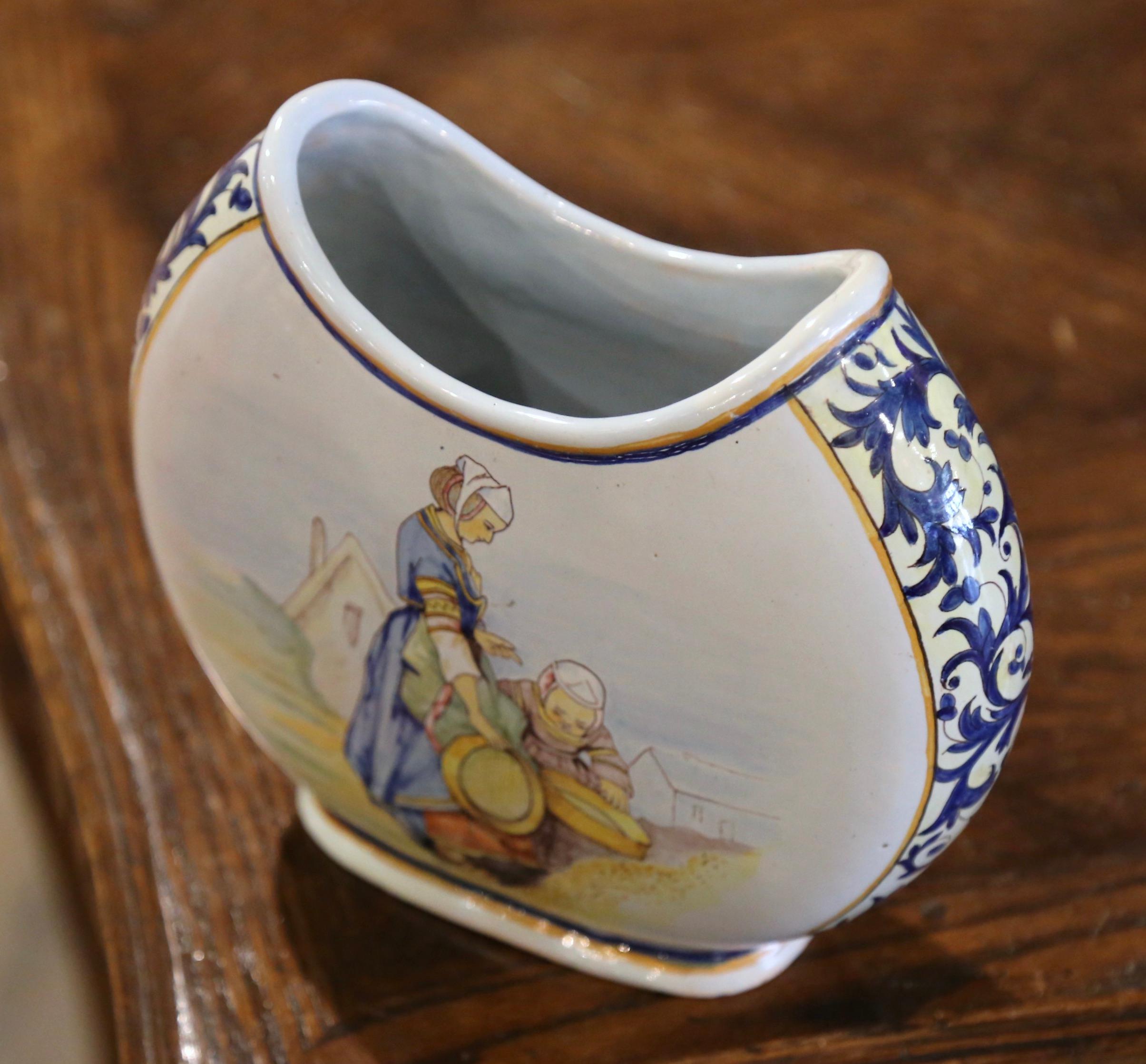 Created in Brittany, France circa 1895, the hand painted faience vase is oval in shape with curved edges at the rim. The vessel is decorated at the center with a pair of Breton ladies sorting out wheat grains in traditional costumes; it is further