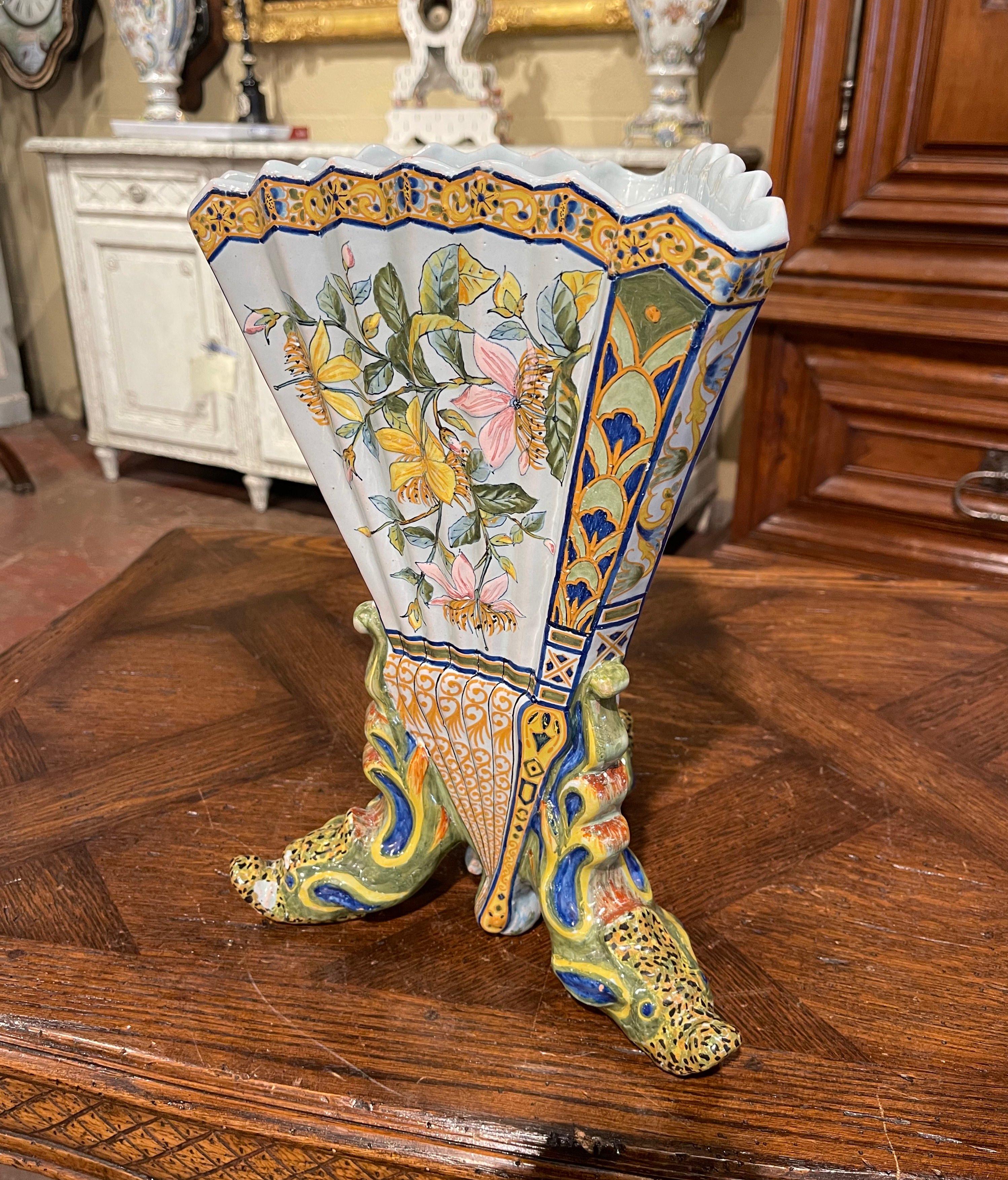Decorate a table or a shelf with this large antique vessel. Created in Brittany, France circa 1895, the colorful hand painted ceramic vase stands on three head shaped feet, over a triangular and tapered form with scalloped rim and accordion shaped