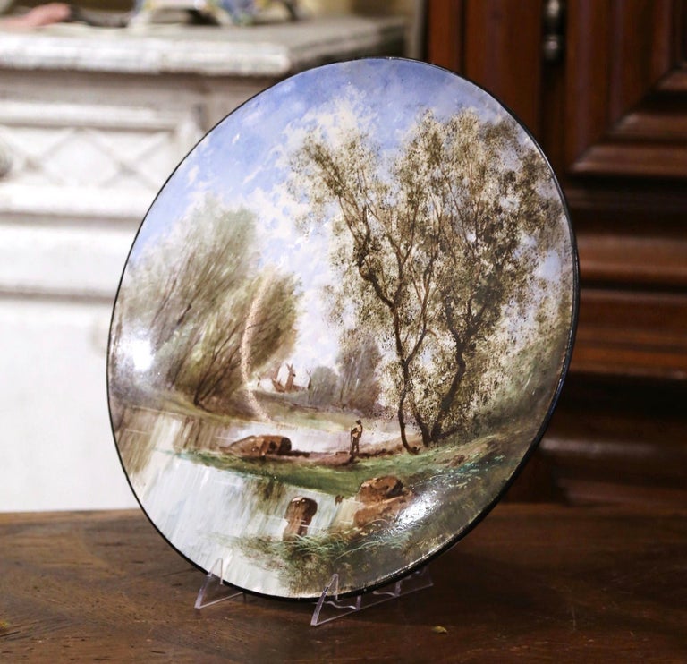 Decorate a wall or a bookcase with this colorful antique wall platter. Crafted in France circa 1890 and round in shape, the large hand painted plate features a pastoral scenery in a luxury landscape; a peasant is shown looking over the pond