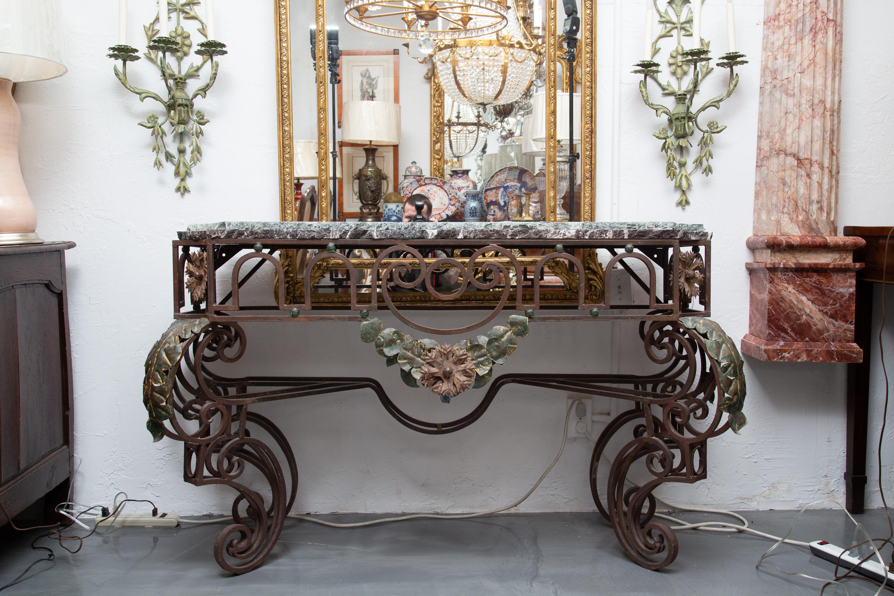 This is an interesting French iron console. The double marble tops with chiseled edge rests over a prominent ornamental frieze with centered polychromed floral spray and supported by out-scrolling intricately forged legs with polychrome leaf