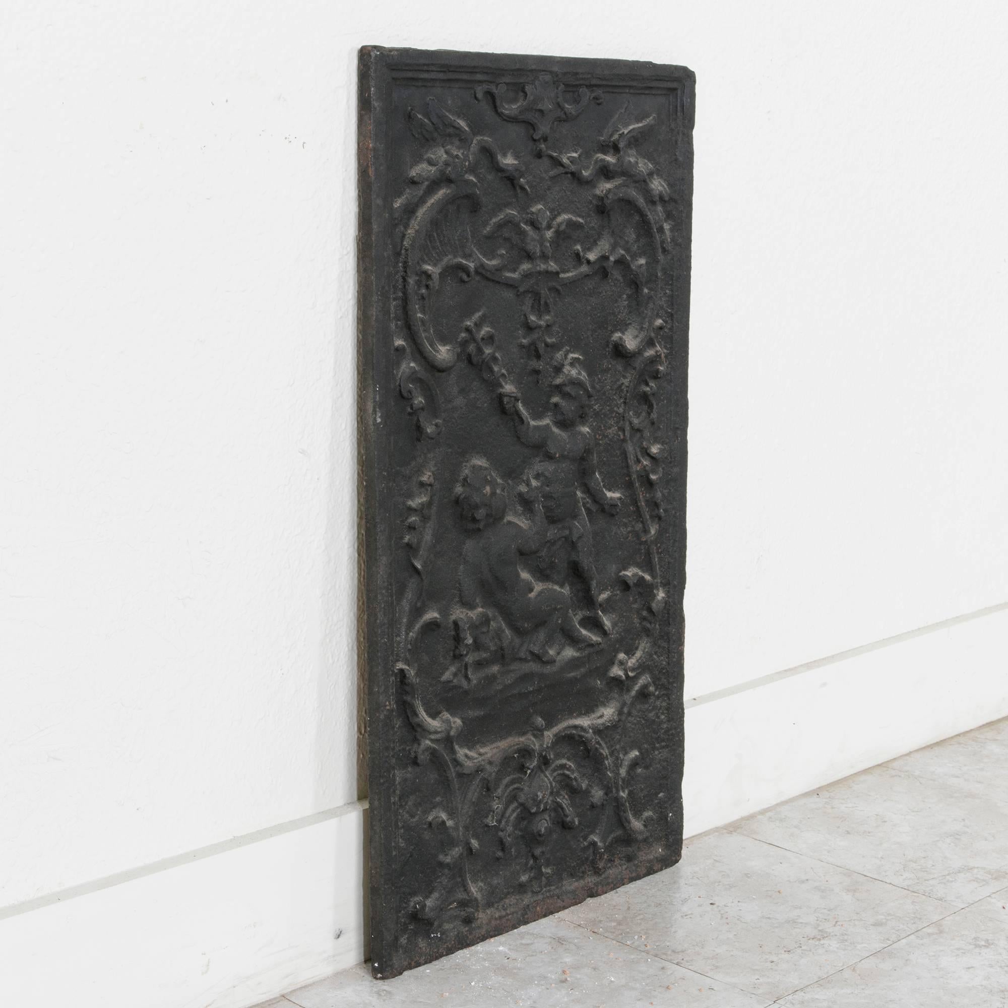 This 19th century iron fireplace back channels a Classic romantic motif featuring putti with scrolling filigree in asymmetrical decoration. One putti carries a staff with two serpents intertwined. This depiction of the caduceus is a reference to the