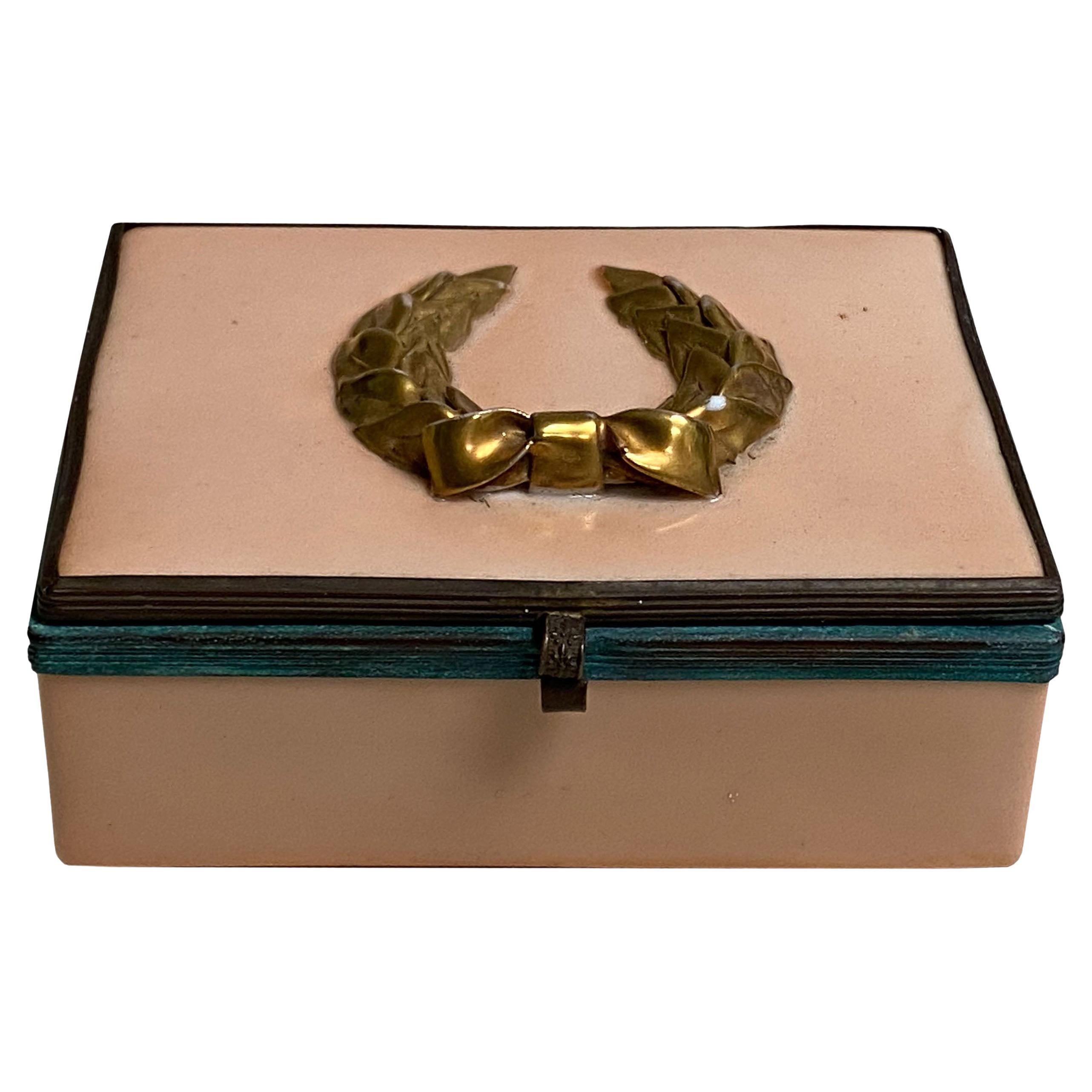 A salmon colored enamel box with patina bronze surround and latch and topped with a gold laurel wreath.