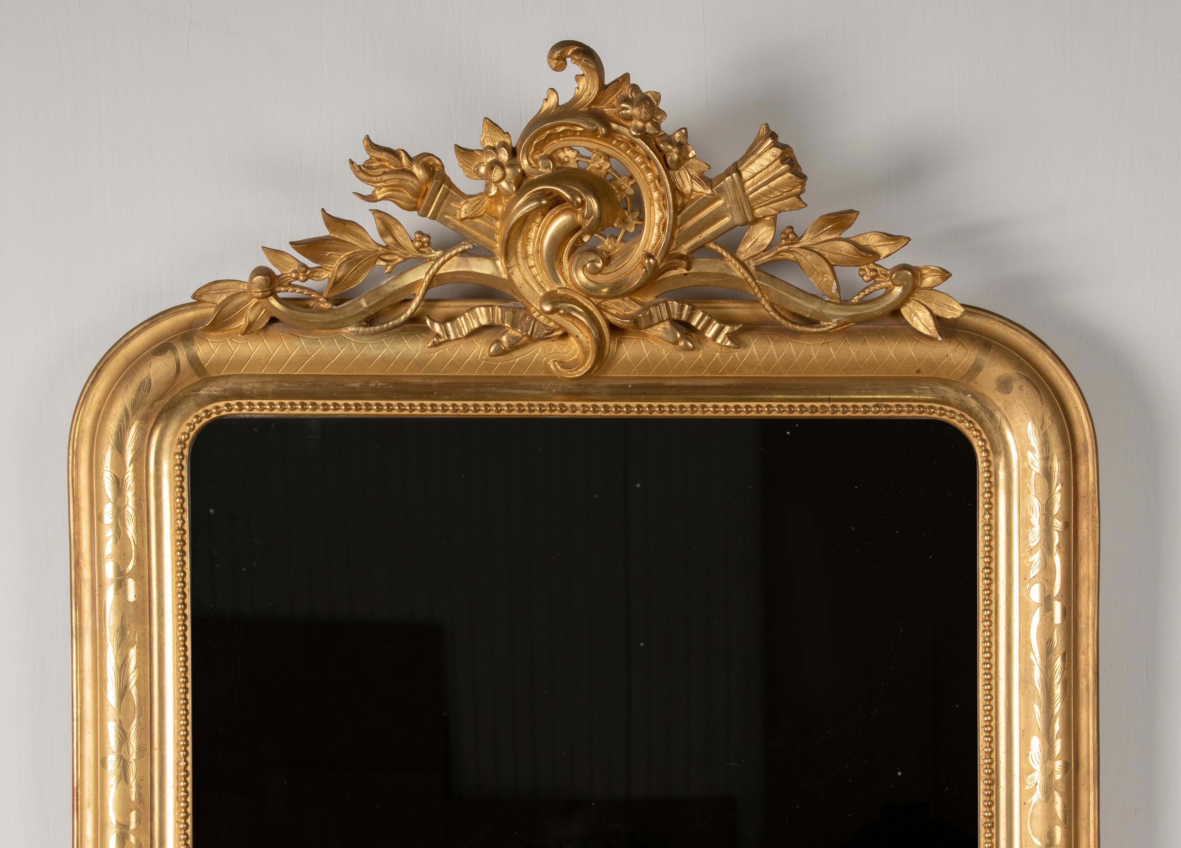 A 19th century Louis Philippe style wall mirror. It has the original gold leaf gilding. On top a crest, the center scrolls are embellished with a bow and arrow, a torch and laurel leaves. The moulded frame rim have a floral etching. The gilding is