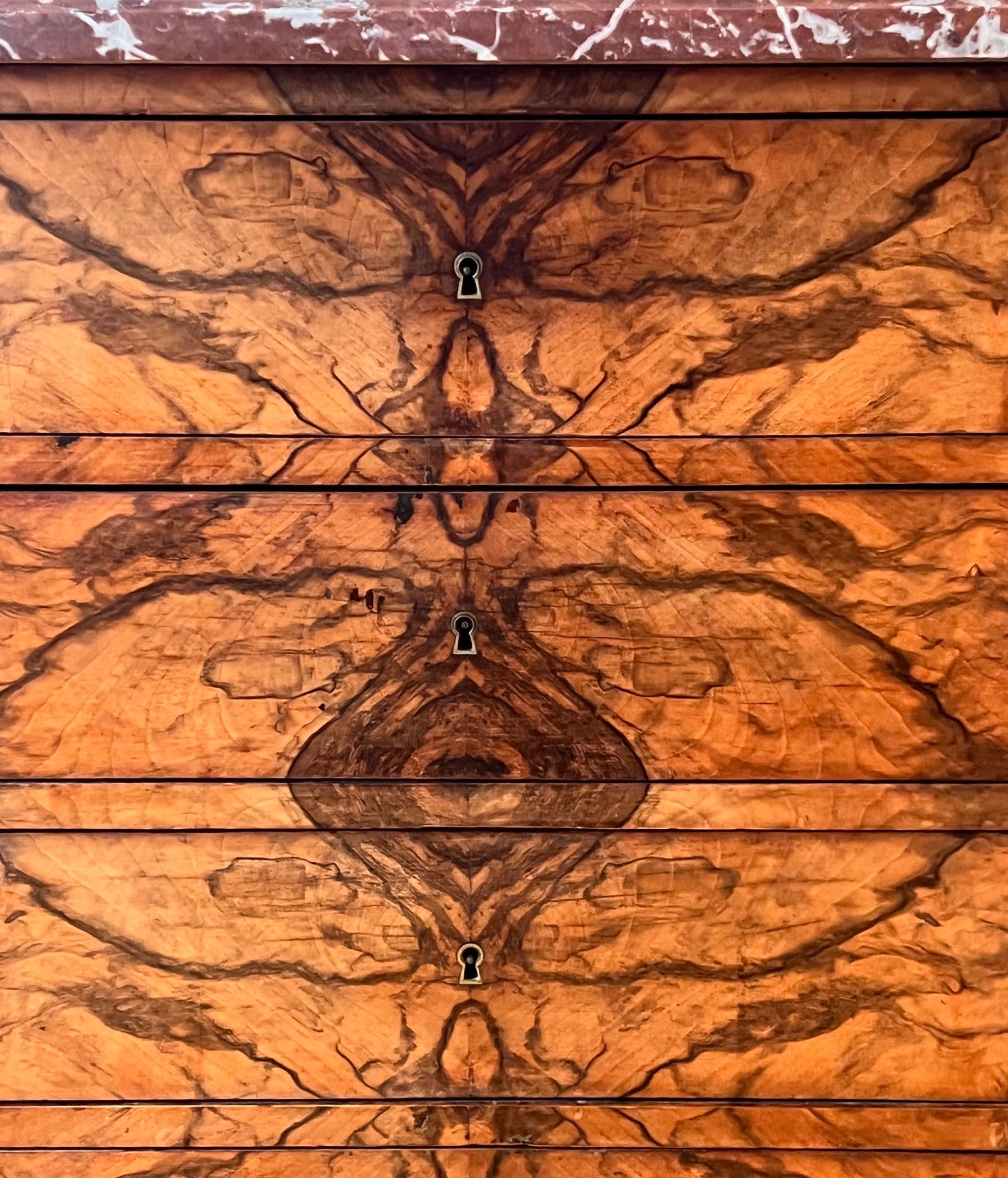 Antique French commode in noce intagliato a mano e accoppiato a libro, con un motivo a specchio simile a una fiamma sui frontali dei cassetti e bordi frontali smussati. Il piano in marmo Rouge-Royal ha gli angoli anteriori arrotondati con venature