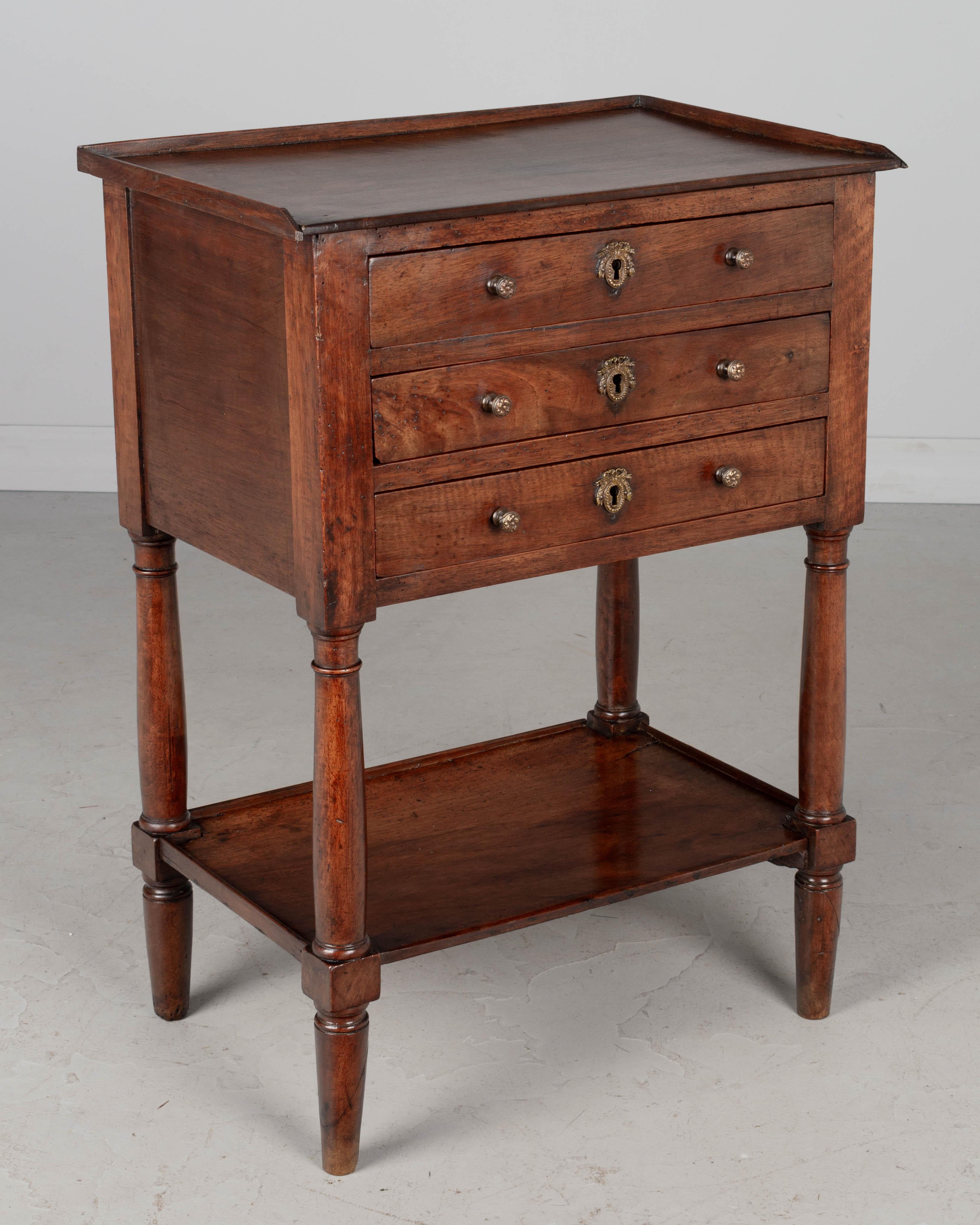 An early 19th century French Louis Philippe walnut side table with three dovetailed drawers, turned legs and lower shelf. Finished on all four sides.  Small brass knobs and decorative escutcheons. Locks are present but no key. Oak as a secondary