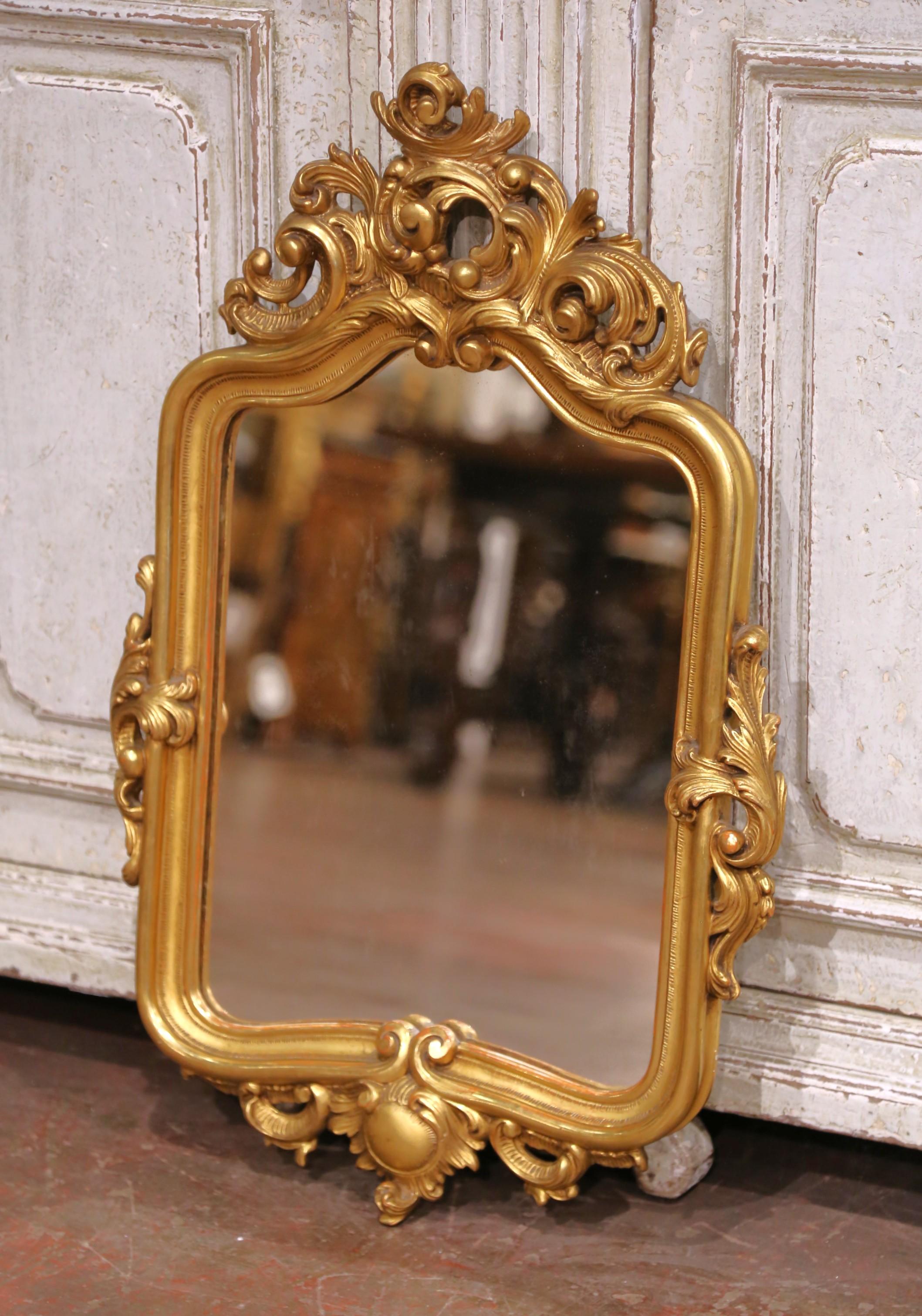 Este encantador espejo de pared antiguo se fabricó en Provenza, Francia, hacia 1880, en estilo Luis XV, conocido por sus elegantes curvas y sus detalles ornamentados. El marco de madera dorada tallada presenta volutas ricamente esculpidas, motivos