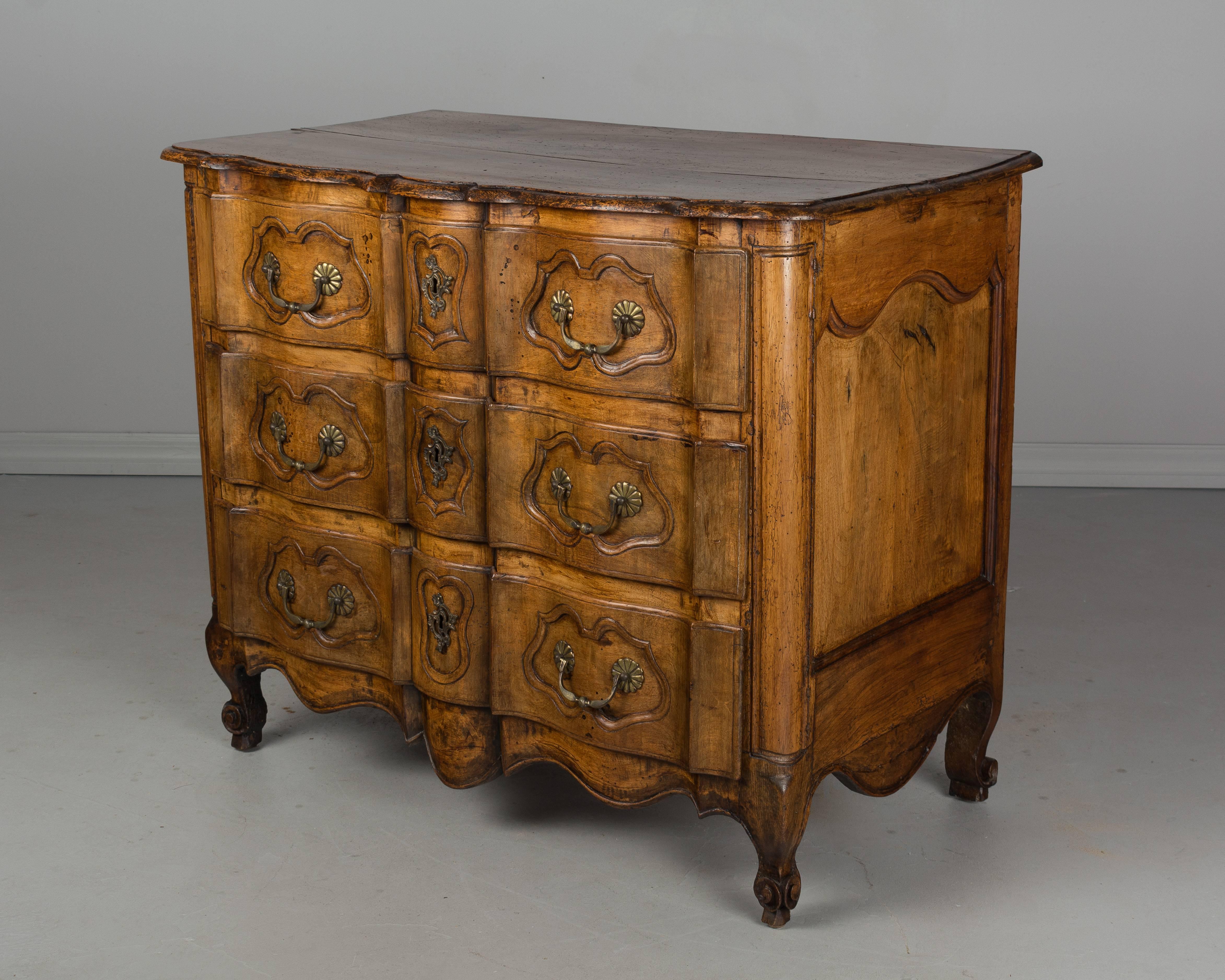 A 19th century French Louis XV style commode with an arbalete, or crossbow, front made of walnut. Three dovetailed drawers with carved details. Brass hardware is of the period, but not original to the piece. Waxed patina. Front legs are replaced. A