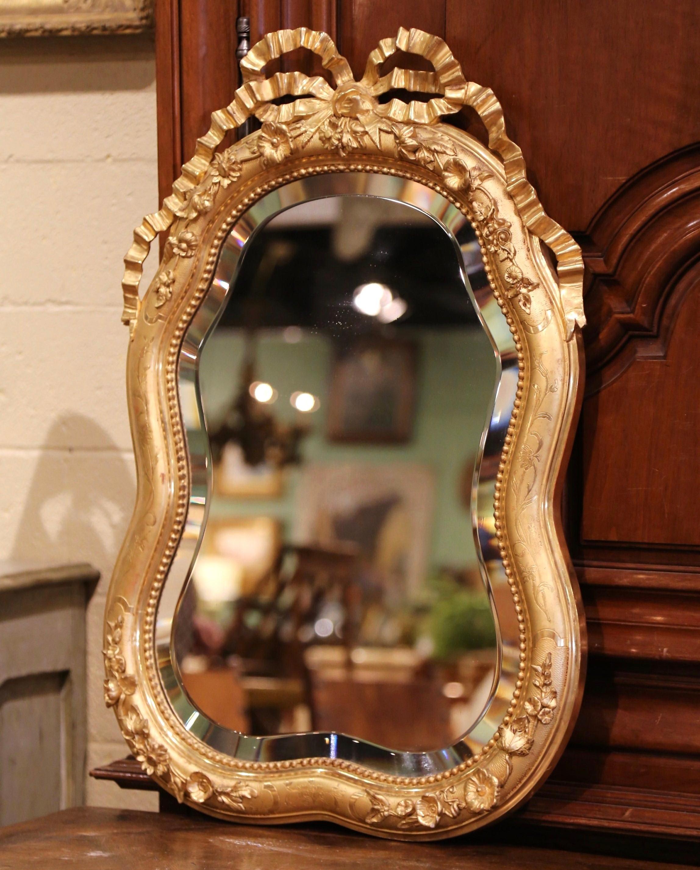 This antique mirror was crafted in Paris, France, circa 1880. Scalloped in shape, the elegant wall hanging mirror features the traditional carved Louis XVI ribbon bow at the pediment with flowers in high relief and engraved floral decor around the