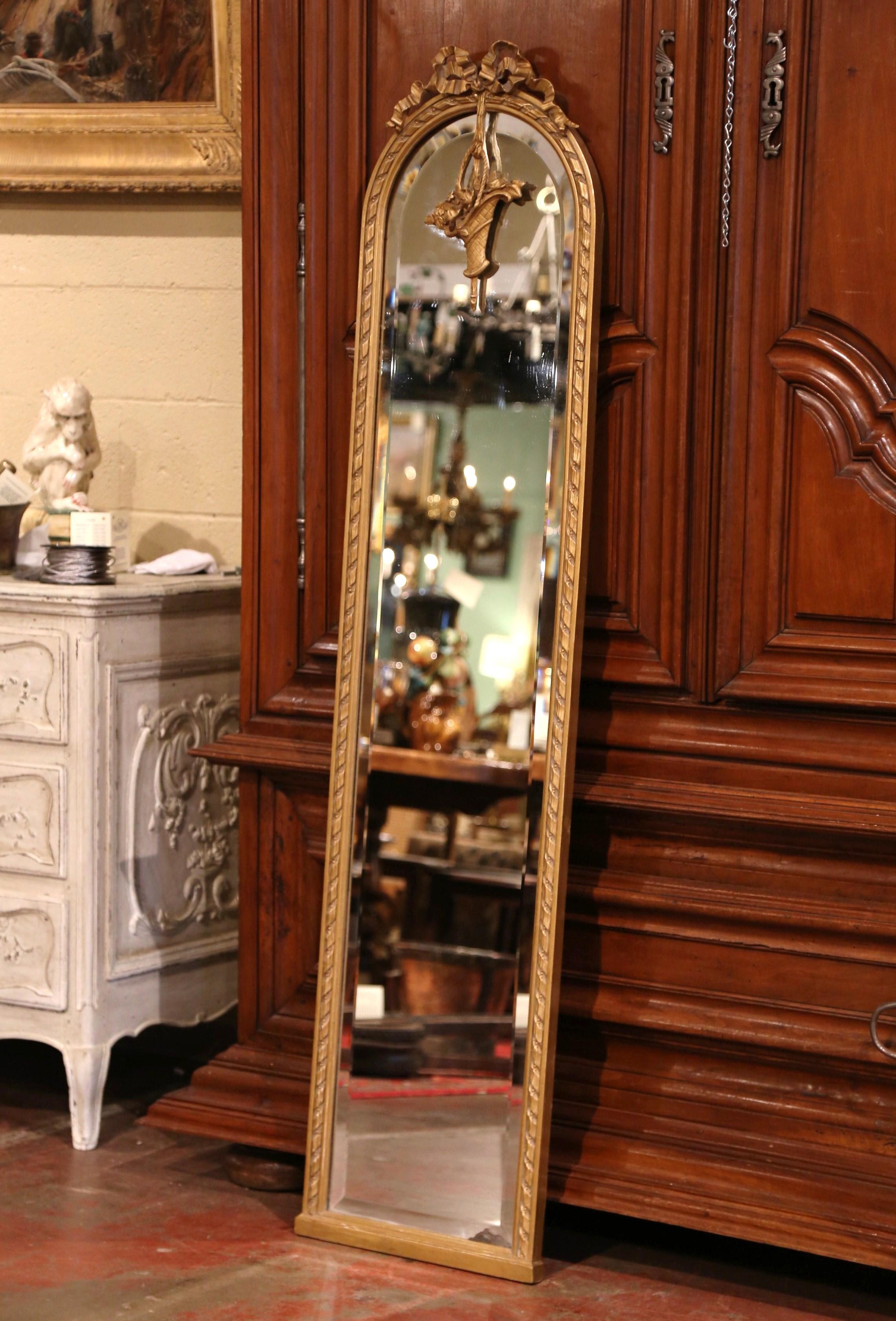 This narrow antique floor mirror was crafted in France, circa 1870. The tall mirror with arched top, features the traditional Louis XVI ribbon bow at the pediment, and is further embellished with a decorative carved basket of flowers hanging down