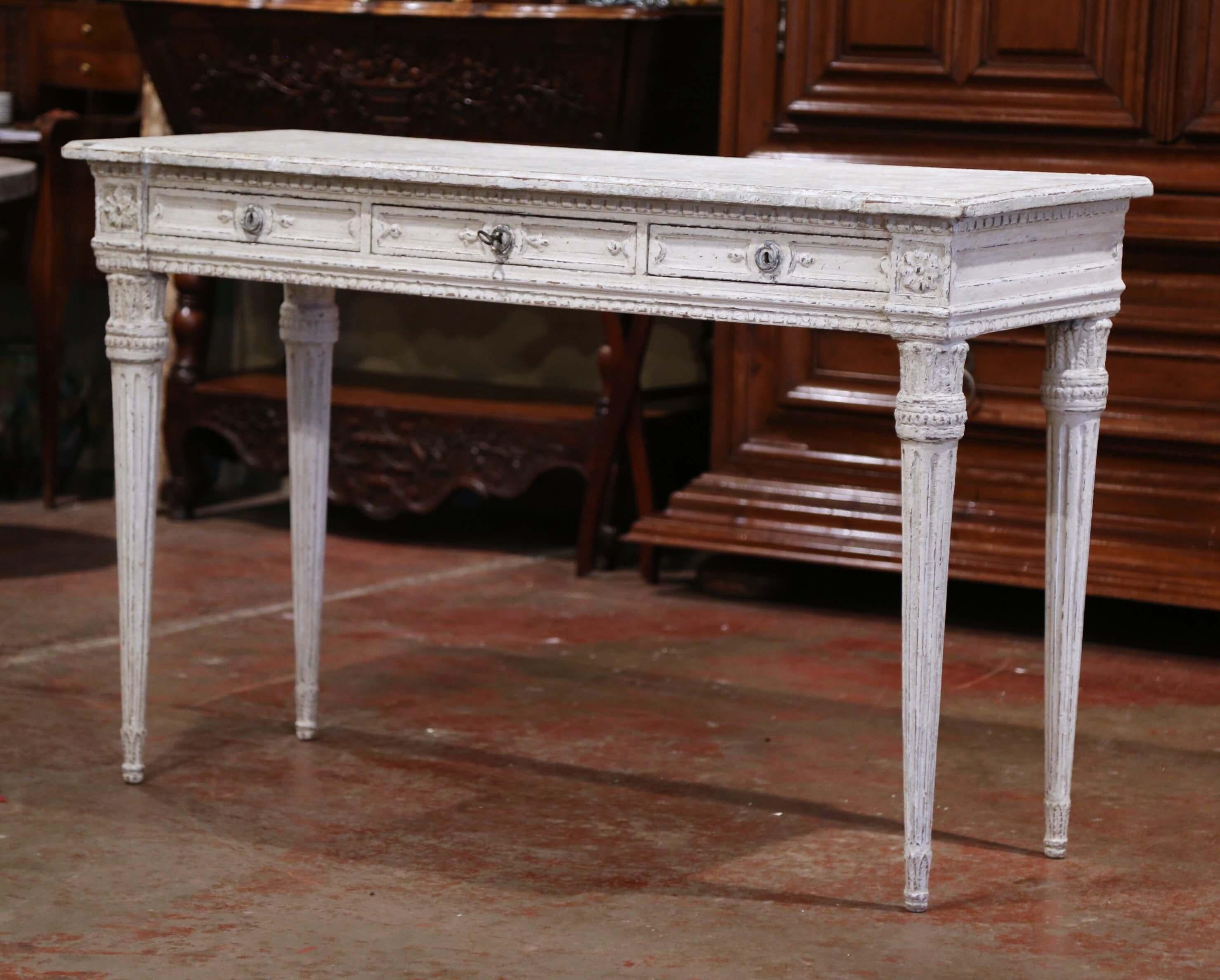 This elegant antique console was created in France, circa 1870. The grey painted table sits on carved and tapered legs over a carved apron embellished with floral medallions on each corner. The console features three drawers across the front dressed