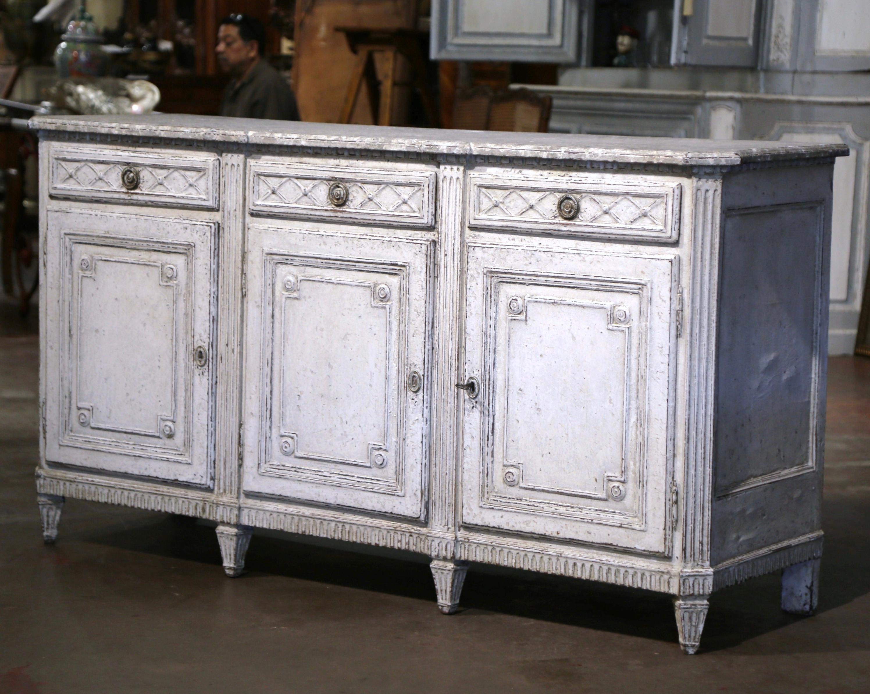 Crafted in France circa 1870, the antique grey painted sideboard stands on eight turned and tapered legs over a straight and carved plinth apron. The traditional enfilade features three doors across the front; each door with inset panel, is