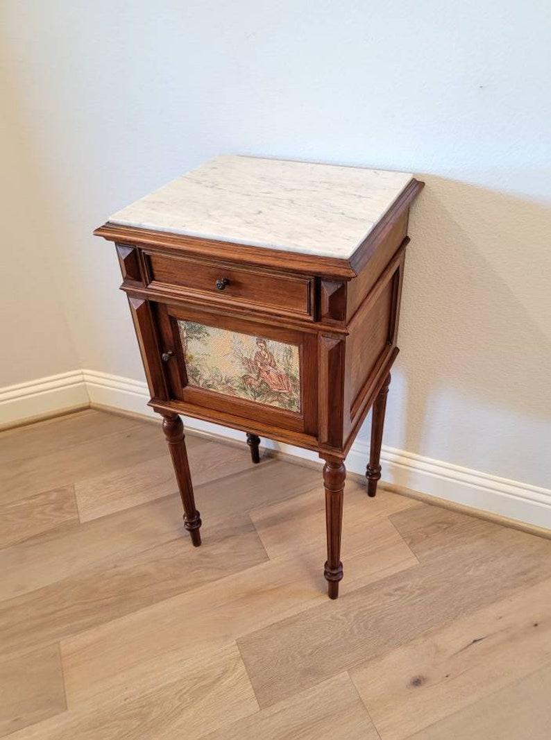 An elegant French antique, circa 1890, bedside cabinet with rare tapestry front. 

Handcrafted in luxurious Louis XVI taste, having a partially inset square gray veined white marble top, framed by a carved stepped molded edge, above a frieze