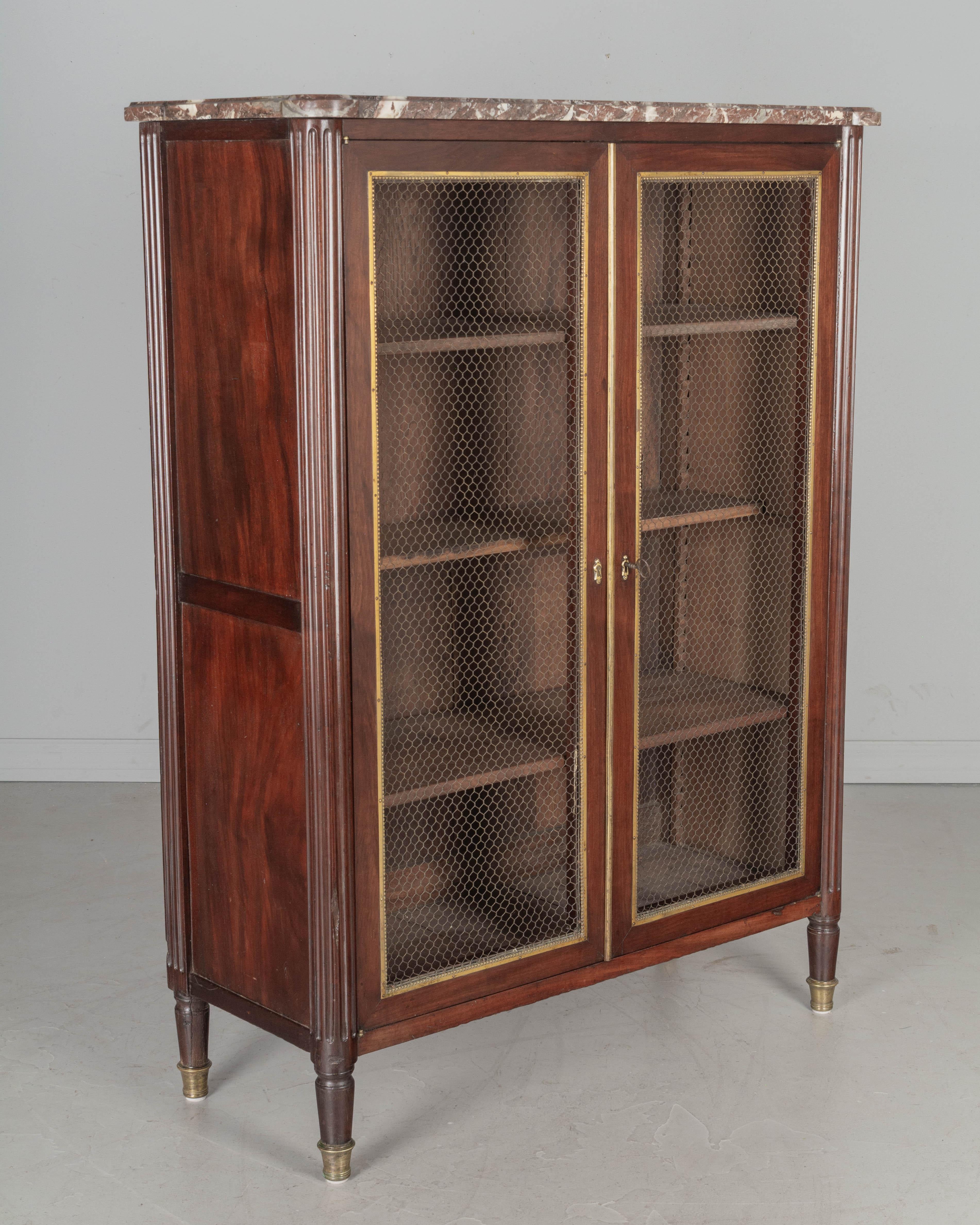 An early 19th Century French Louis XVI style marble top bibliotheque, or bookcase. Made of solid mahogany and retaining a nice high gloss luster finish. Brass grillage doors with decorative beaded brass trim, working lock and key. Rounded fluted