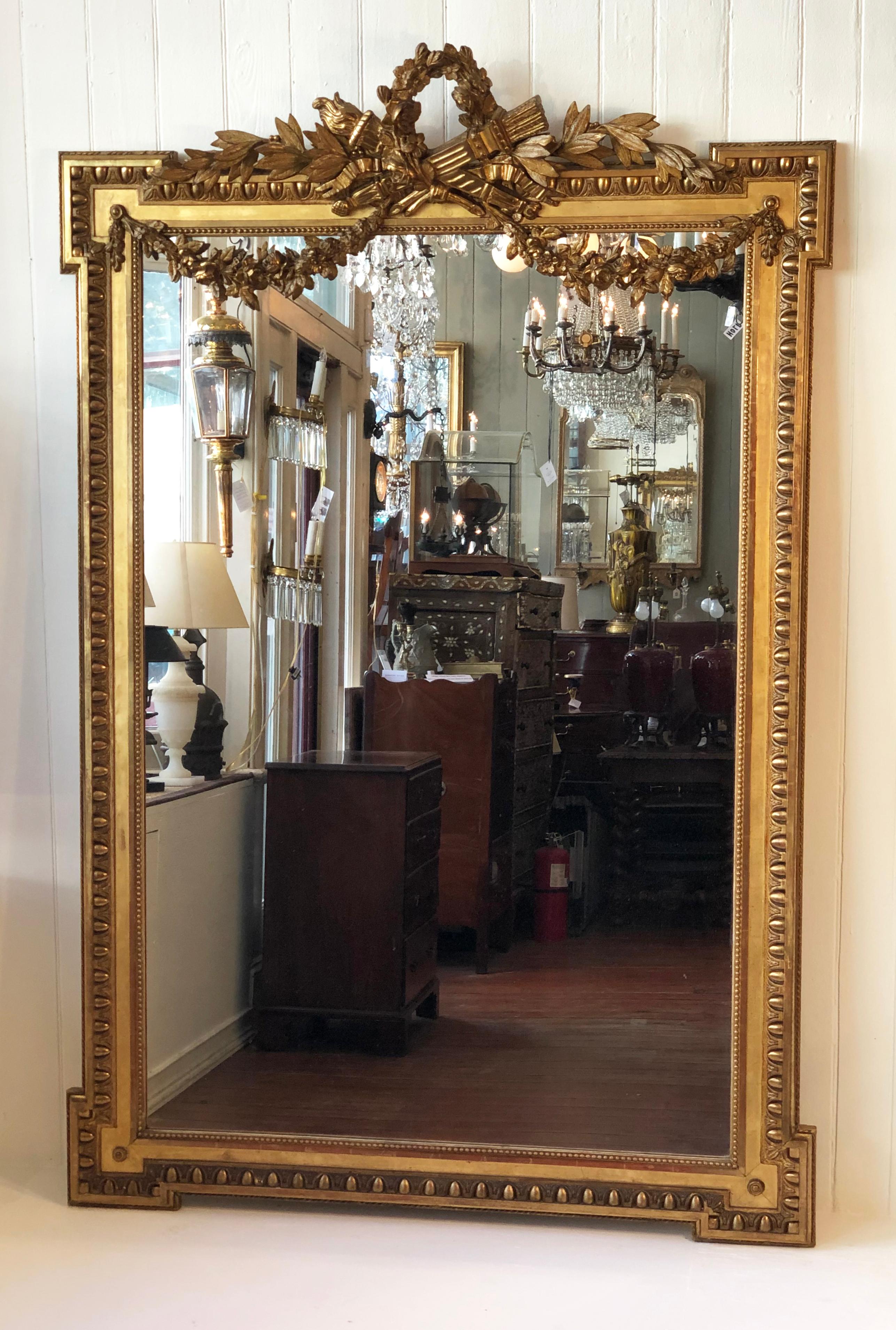 Grand 19th century French Louis XVI style gilded mirror, circa 1840-1860. This wonderful mirror is adorned with a floral wreath cartouche, also having crossed flambeau and quiver in the center of the cartouche above garland swags. The frame has four