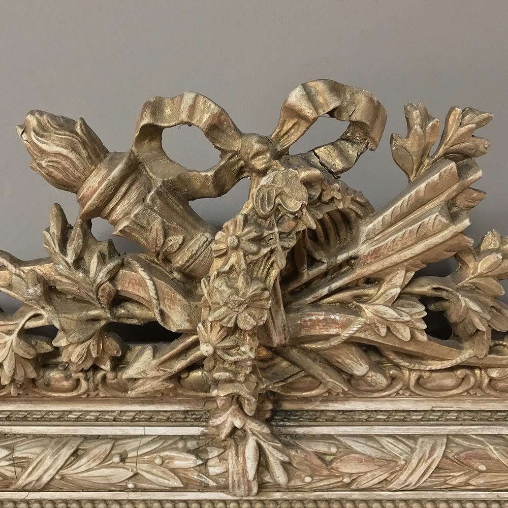 19th century French Louis XVI gilded mirror boasts an elaborately detailed crown depicting the regal torchère and carquois de fleche intertwined with an Archer's bow, ribbon, and laurels with foliates. Laurel wreath punctuated with acanthus rosettes